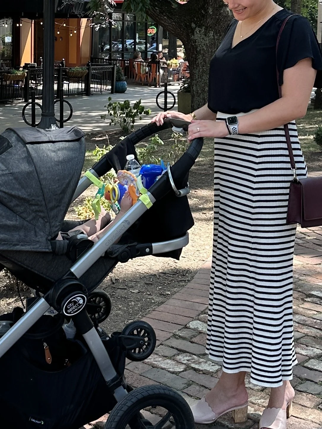 A woman in a black top and striped skirt is pushing a stroller containing a baby with toys and a water bottle, on a brick sidewalk in an outdoor area with trees and a restaurant or café in the background.