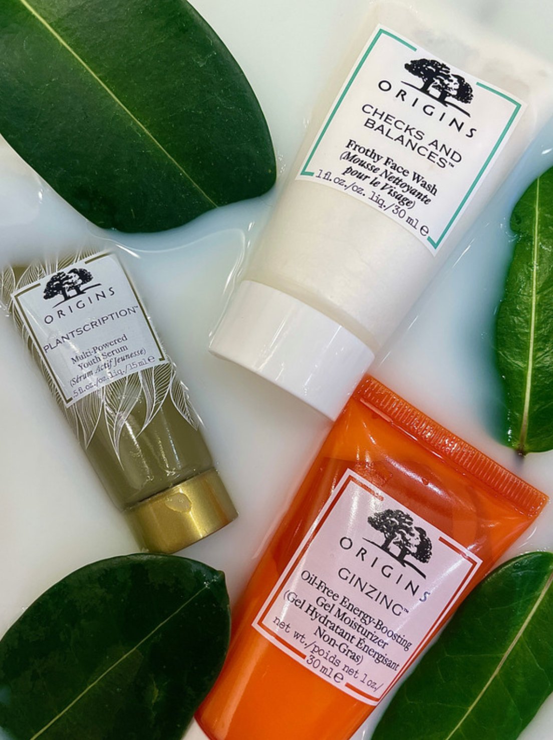 Skincare products from Origins, including Frothy Face Wash, Multi-Powered Youth Serum, and Oil-Free Energy-Boosting Gel Moisturizer, arranged on a white surface with green leaves.