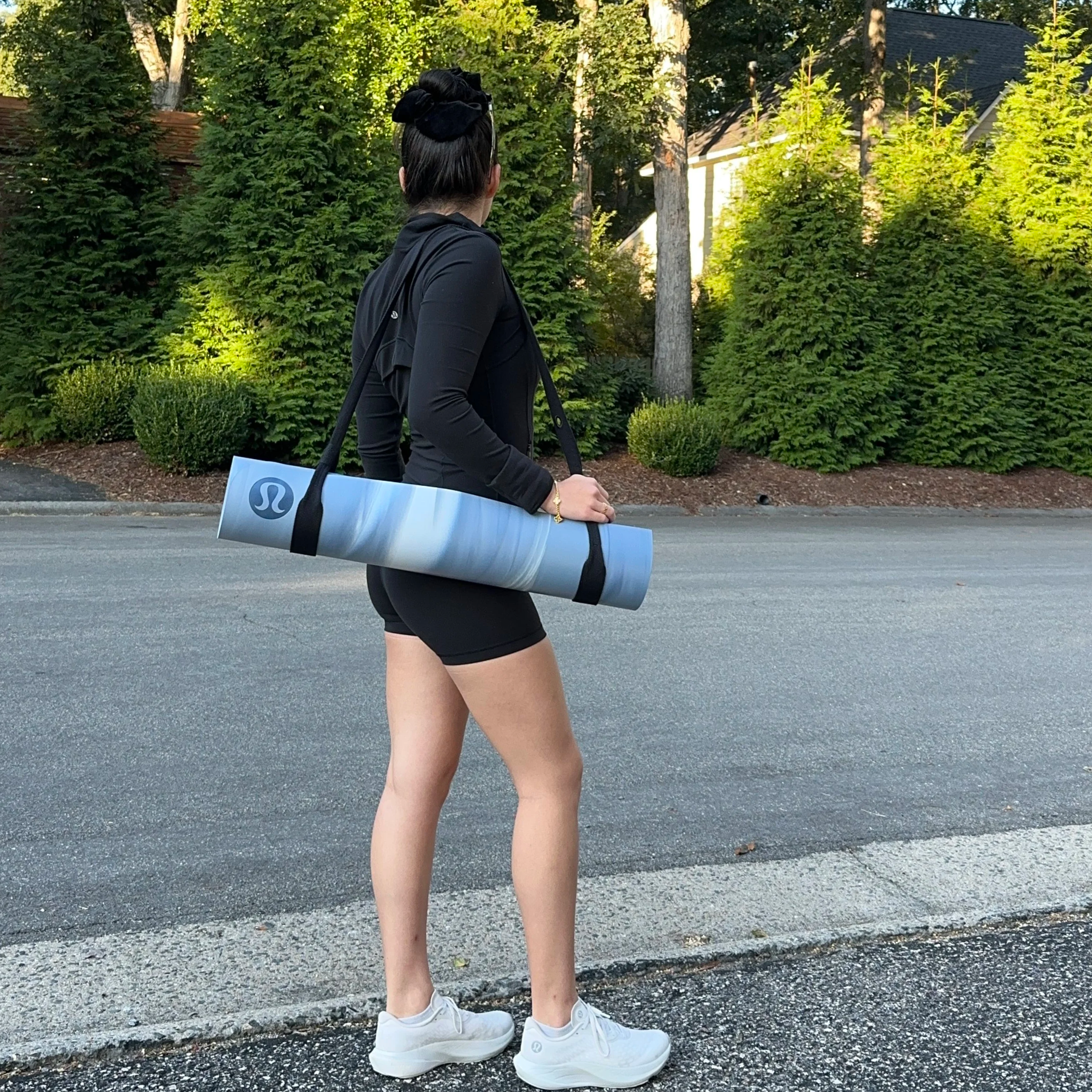 A woman dressed in black sportswear holding a yoga mat with a Lululemon logo, standing on a sidewalk with greenery and trees in the background.
