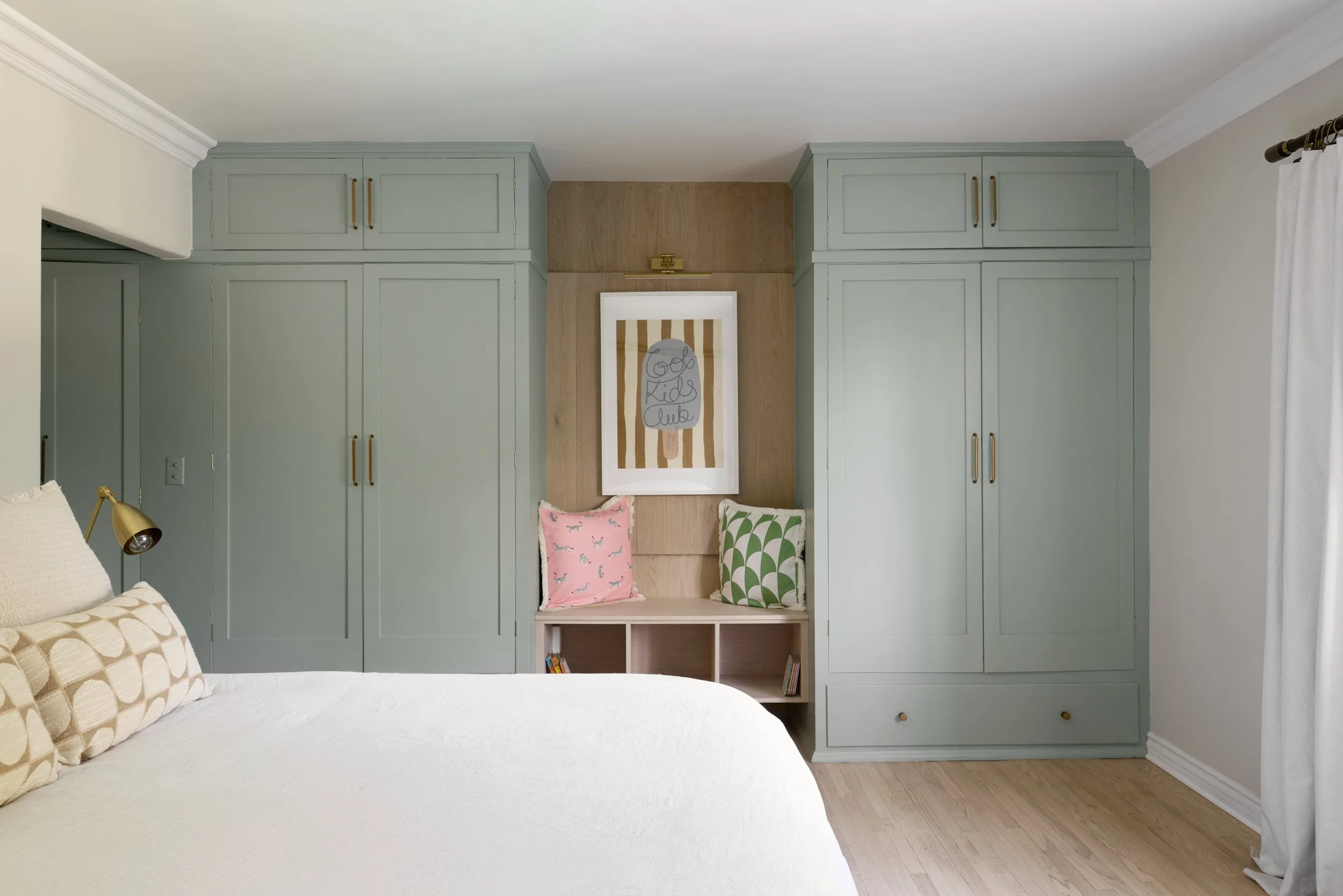 A bedroom with a bed, built-in light green wardrobes, a wall with wood paneling, and a framed art piece with striped background. There are two decorative pillows, one pink with dog illustrations and one green with leaf patterns, on a small bench with cubbies containing books. White curtains are partially visible on the right.