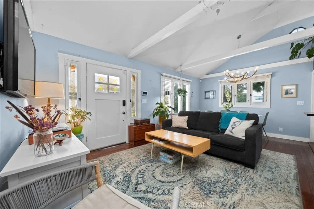 Living room with light blue walls, white ceiling beams, a black sofa with white and turquoise pillows, a wooden coffee table, a large patterned rug, multiple windows, potted plants, and framed pictures.