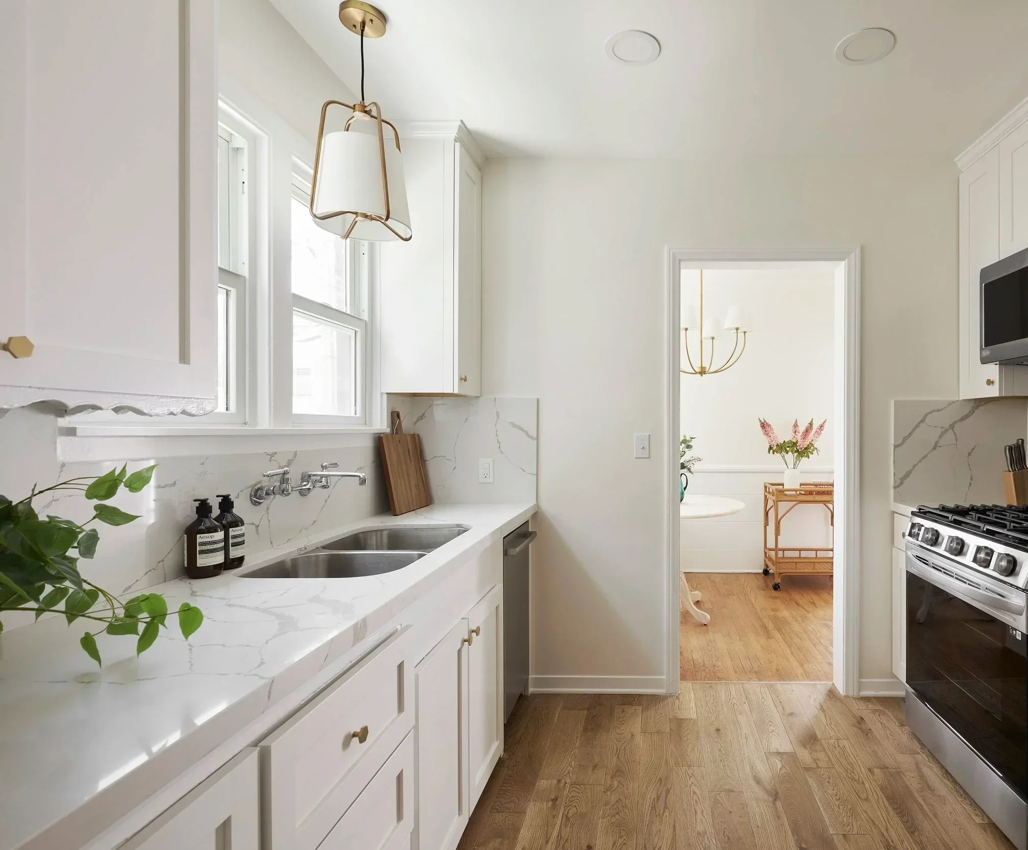 A bright kitchen with white cabinets, a marble countertop, a stainless steel sink, a window, and a modern light fixture. Through the doorway, a dining area with a chandelier and a table with pink flowers is visible.