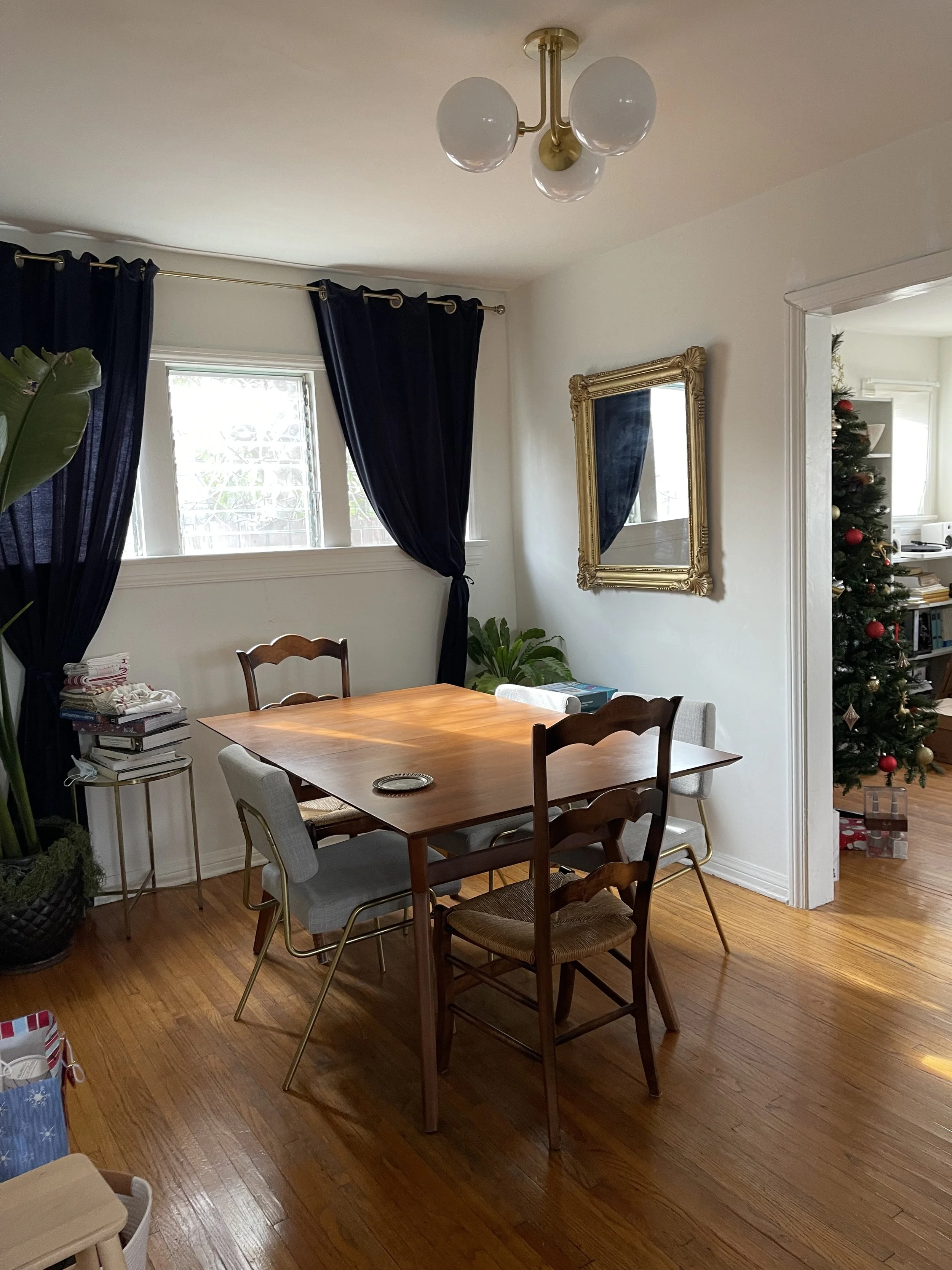 A dining room with a wooden table and mixed chair styles, navy curtains on windows, a gold-framed mirror on the white wall, and a Christmas tree with presents in the background.