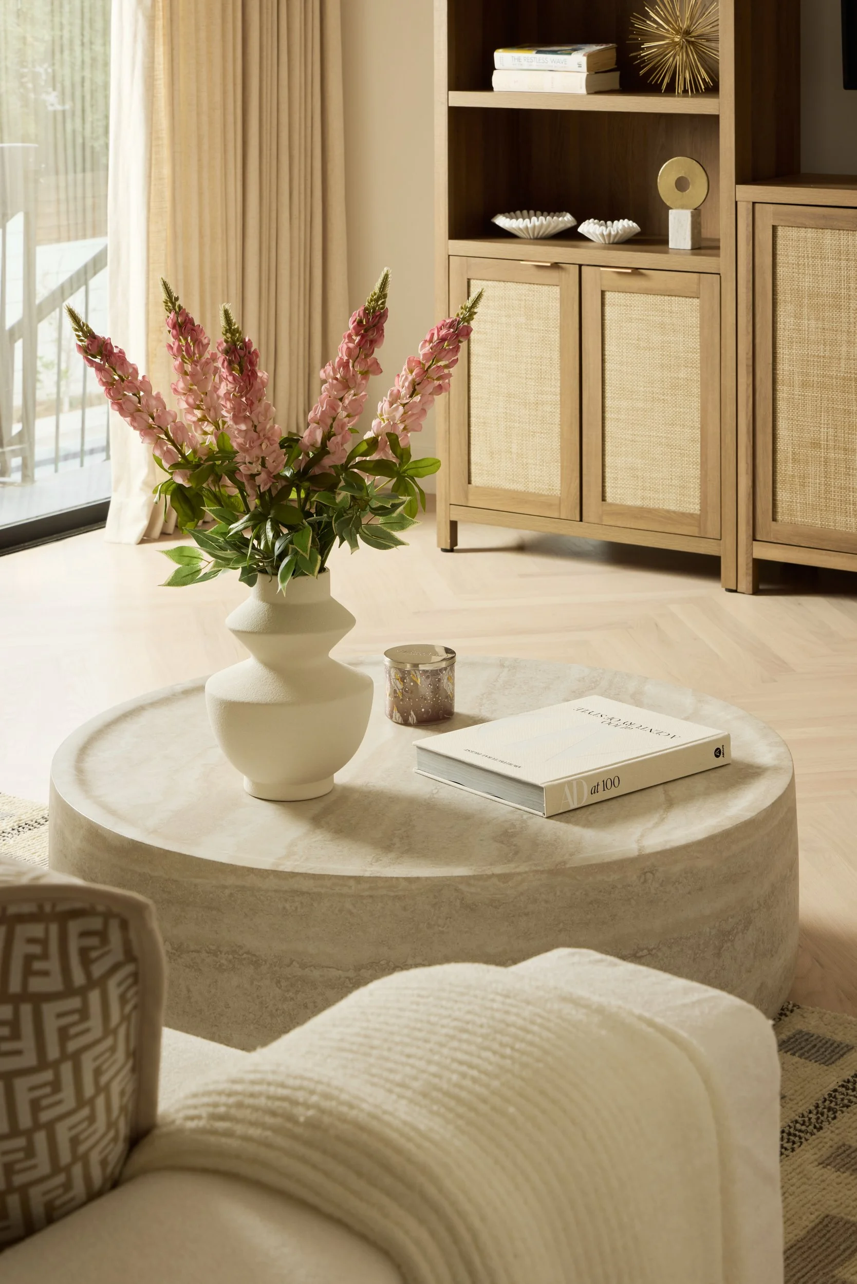 A living room with a round marble coffee table holding a white vase with pink flowers, a candle, and a book. In the background, there is a wooden cabinet with decorative objects, and a sliding glass door with beige curtains.