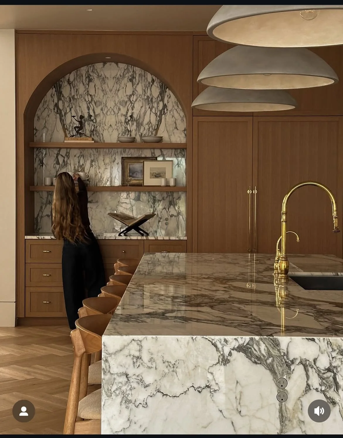 A person with long hair reaching for a cup on a wooden shelf in a kitchen. The kitchen features a marble backsplash, a marble island countertop, and gold fixtures.