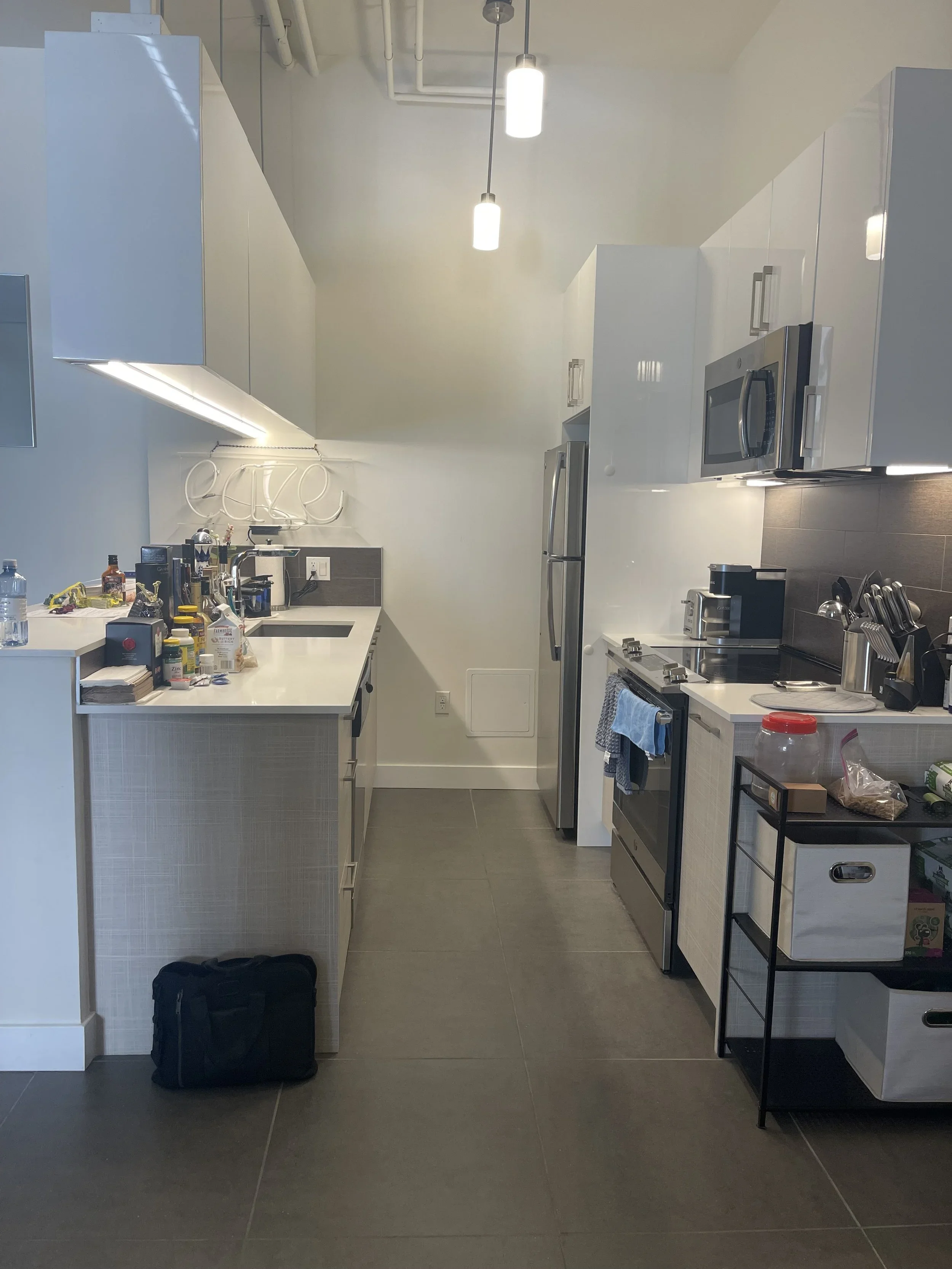 Modern kitchen with white cabinets, stainless steel appliances, and gray tile flooring. Cluttered countertops, hanging pendant lights, and a small rolling cart.