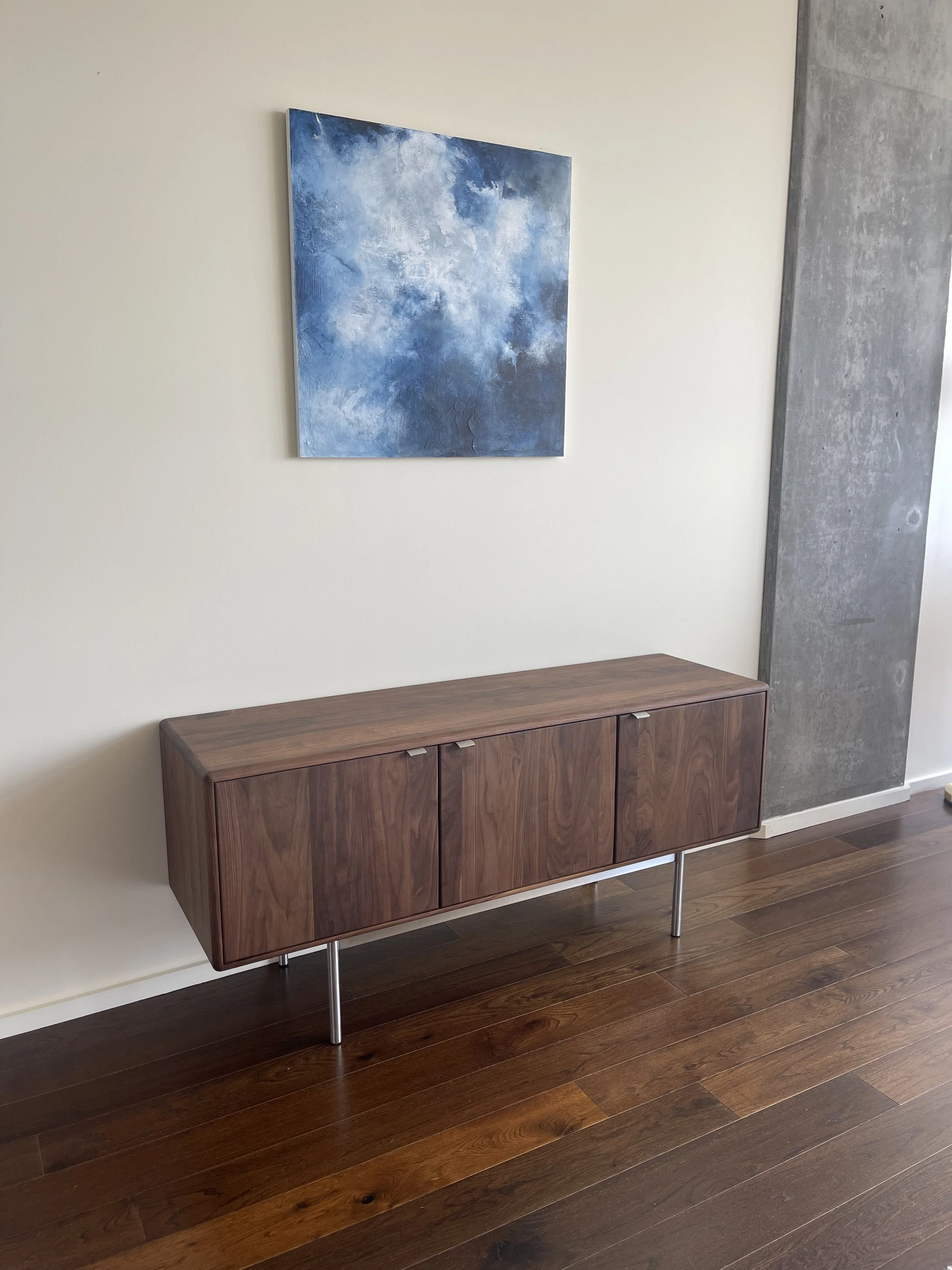 A mid-century modern wooden sideboard with three doors and metal legs, placed against a white wall. Above it hangs an abstract blue and white painting, and the floor is made of polished brown hardwood.