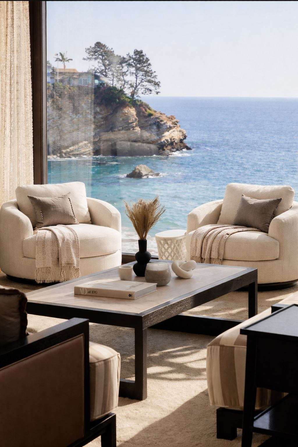 Living room with two beige armchairs, a marble coffee table, and a large window overlooking the ocean and rocky coastline.