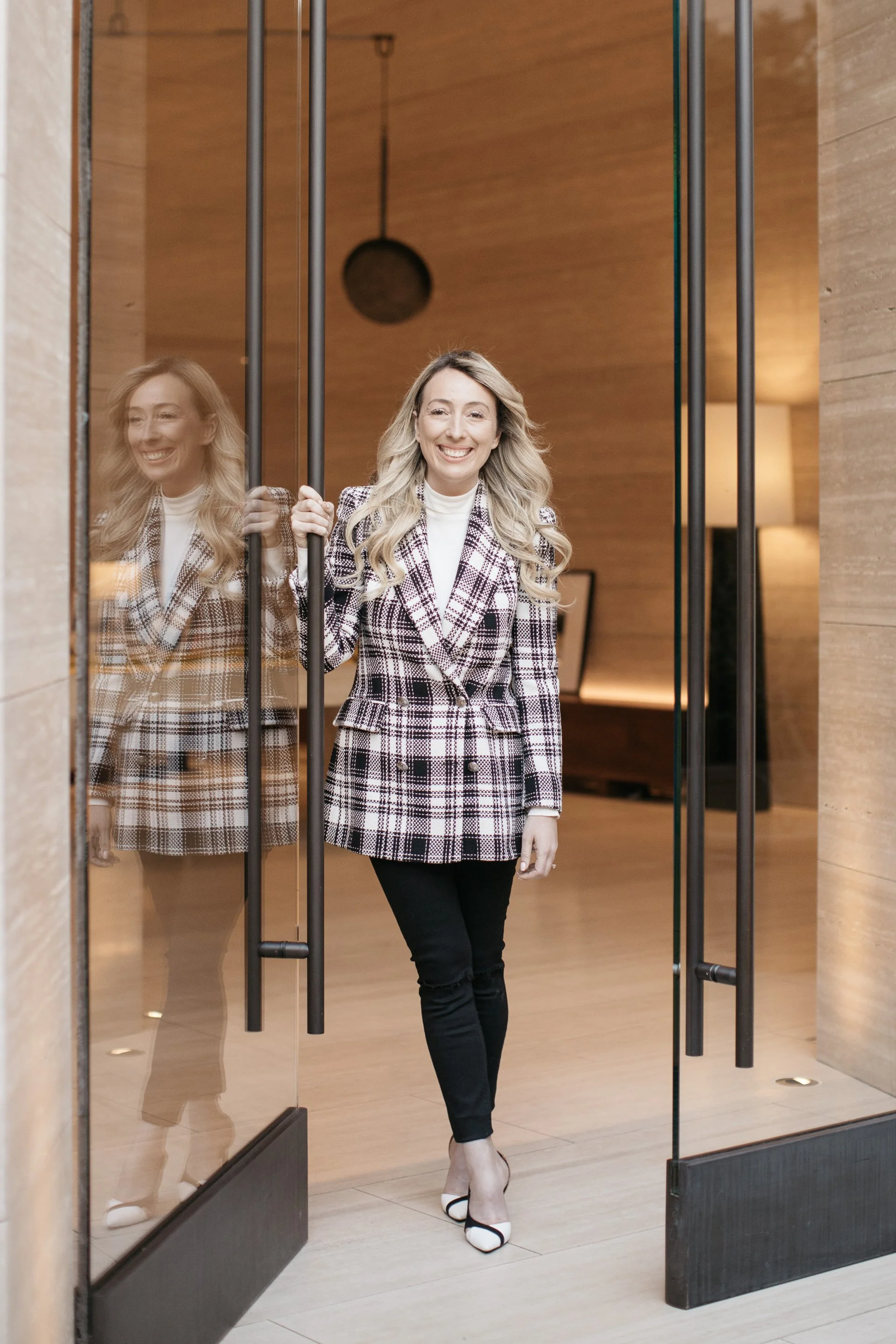 A woman with blonde hair wearing a plaid blazer, white turtleneck, black pants, and white heels stepping through glass doors, smiling.