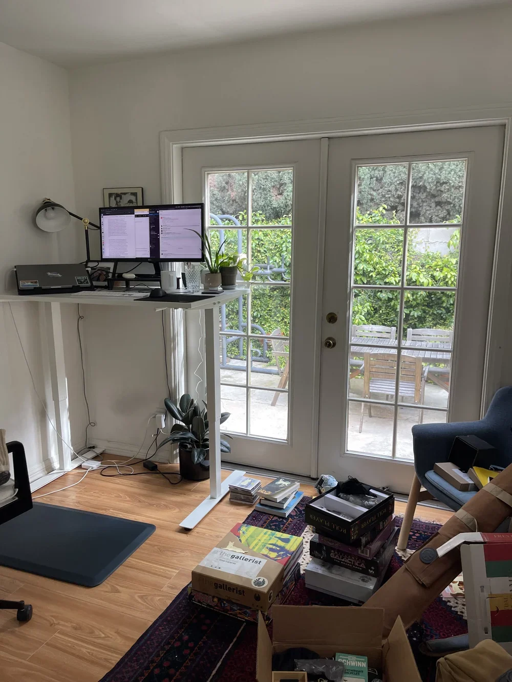 Home office with a laptop on a white standing desk, dual monitors, potted plants, wooden deck outside glass double doors, and scattered board games and books on a colorful rug.