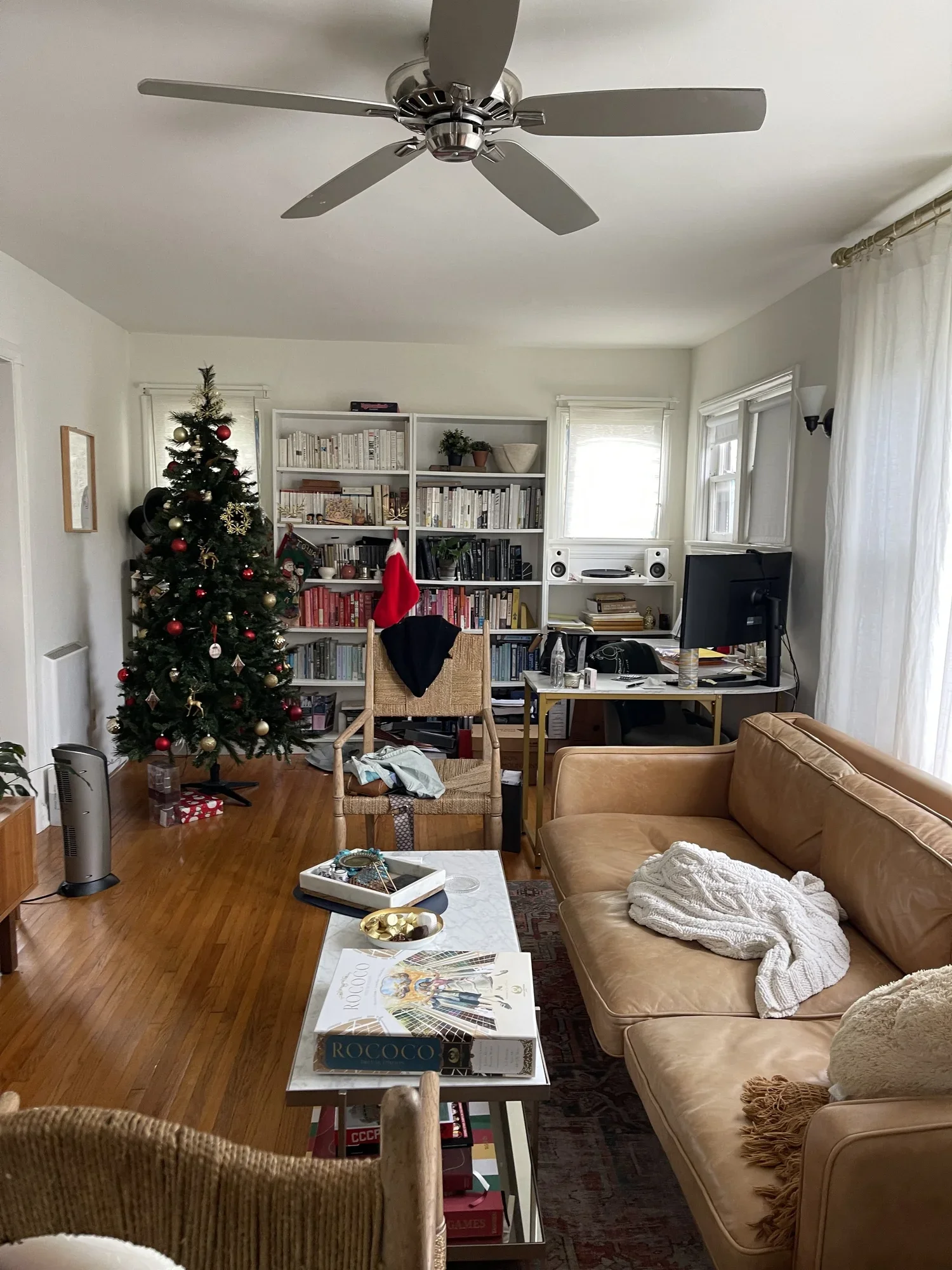 A cozy living room decorated for Christmas with a Christmas tree adorned with red and gold ornaments, a bookshelf filled with books, a tan sofa with a white blanket, a wooden chair with a Santa hat hanging on it, a white coffee table with puzzles and a board game, a computer desk with two monitors, a speaker, and a water bottle, a heater, and a window with sheer curtains.