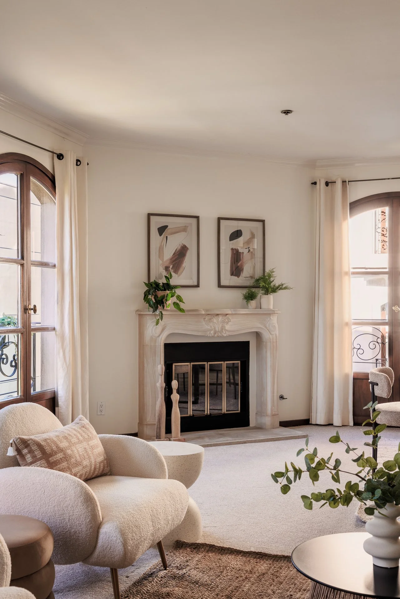 Living room with cream-colored furniture, a fireplace with a carved mantel, framed abstract art above the mantel, potted plants, and large windows with white curtains.