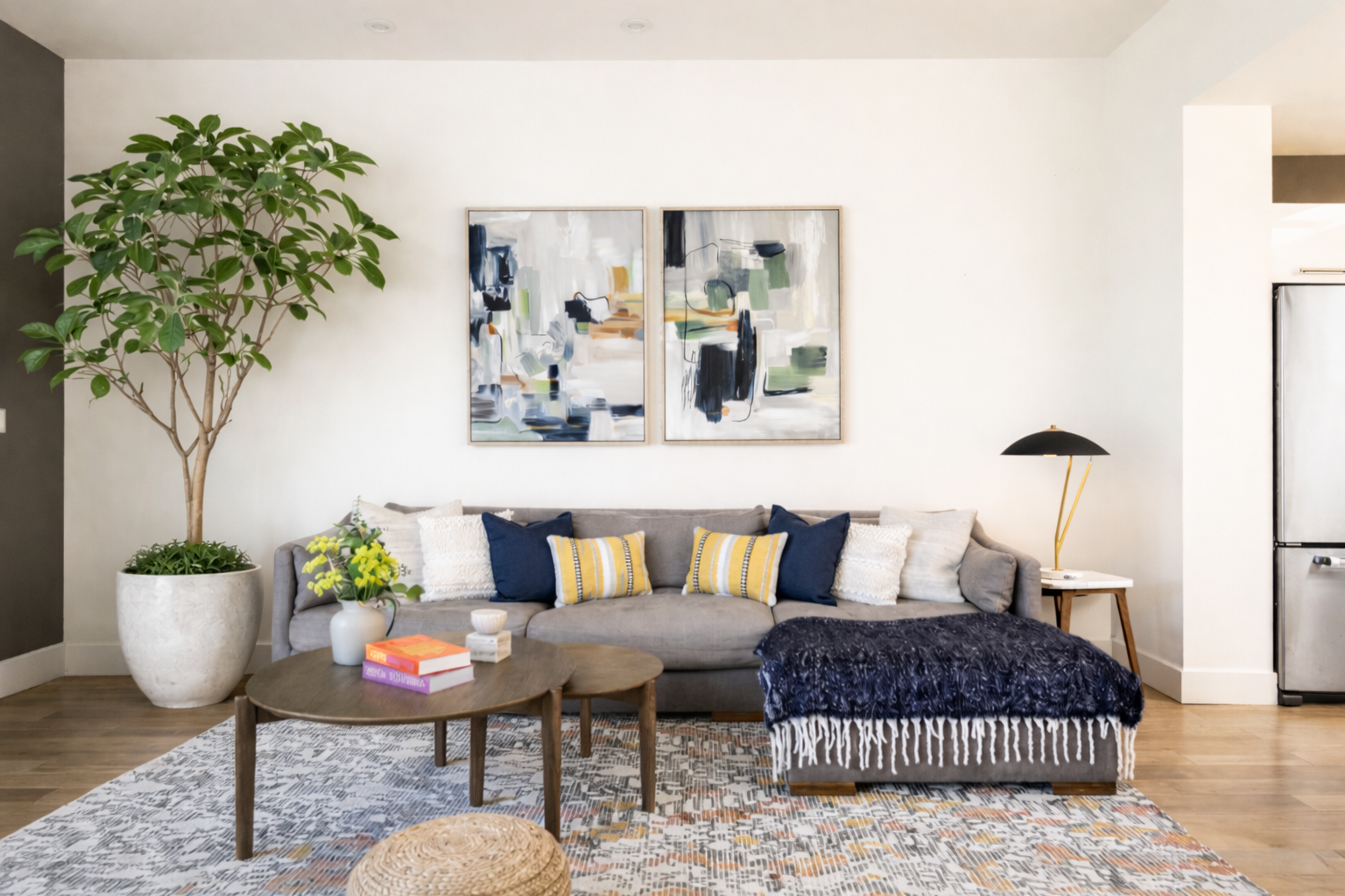 Living room with gray sofa, blue and yellow pillows, abstract paintings, large potted plant, coffee table with books and a vase, patterned rug, black and gold floor lamp, and a stainless steel refrigerator.