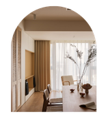 Modern dining room with a wooden table, six chairs, and a white vase with branches. Large windows with curtains allowing natural light, and built-in storage cabinetry.