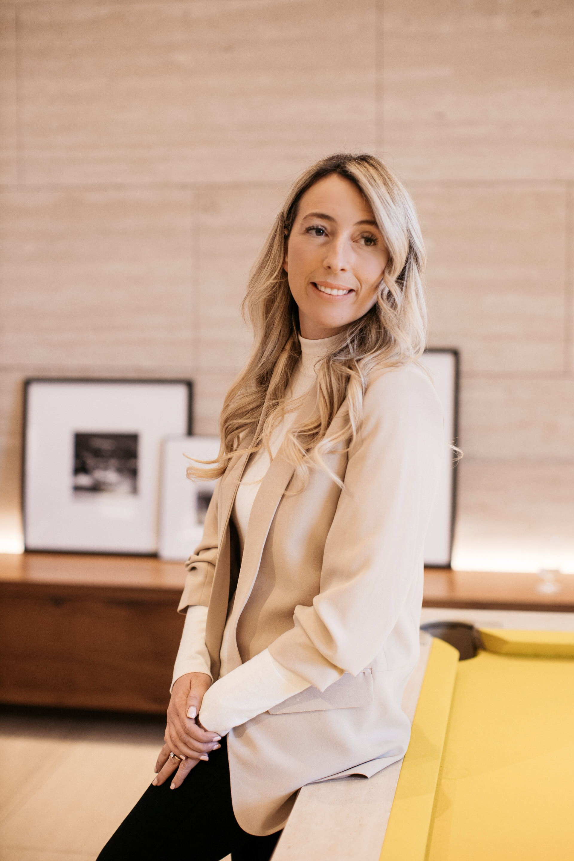 A woman with blonde, wavy hair sitting on a yellow bench in an office or gallery space, wearing a beige blazer and a white top, smiling at the camera with black framed photographs on a wooden surface behind her.