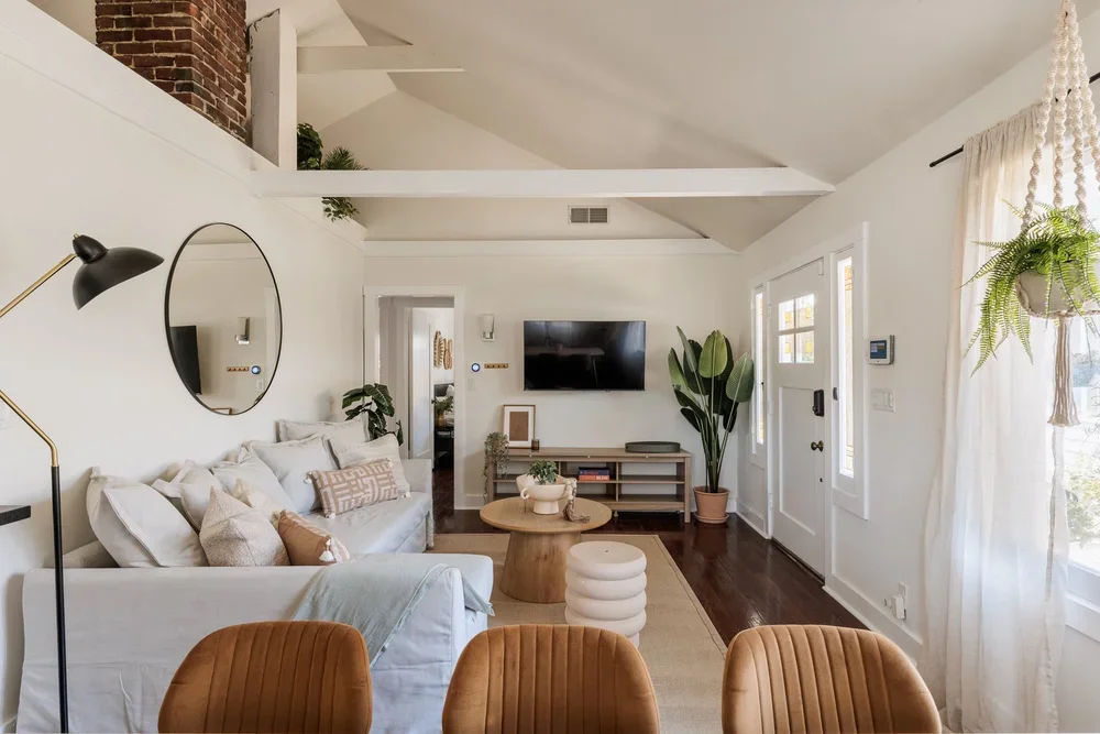 Living room with white walls, a white sofa with beige and white pillows, a black floor lamp, a round mirror, a flat-screen TV on the wall, large green plants, a wooden coffee table, and curtains on the window.