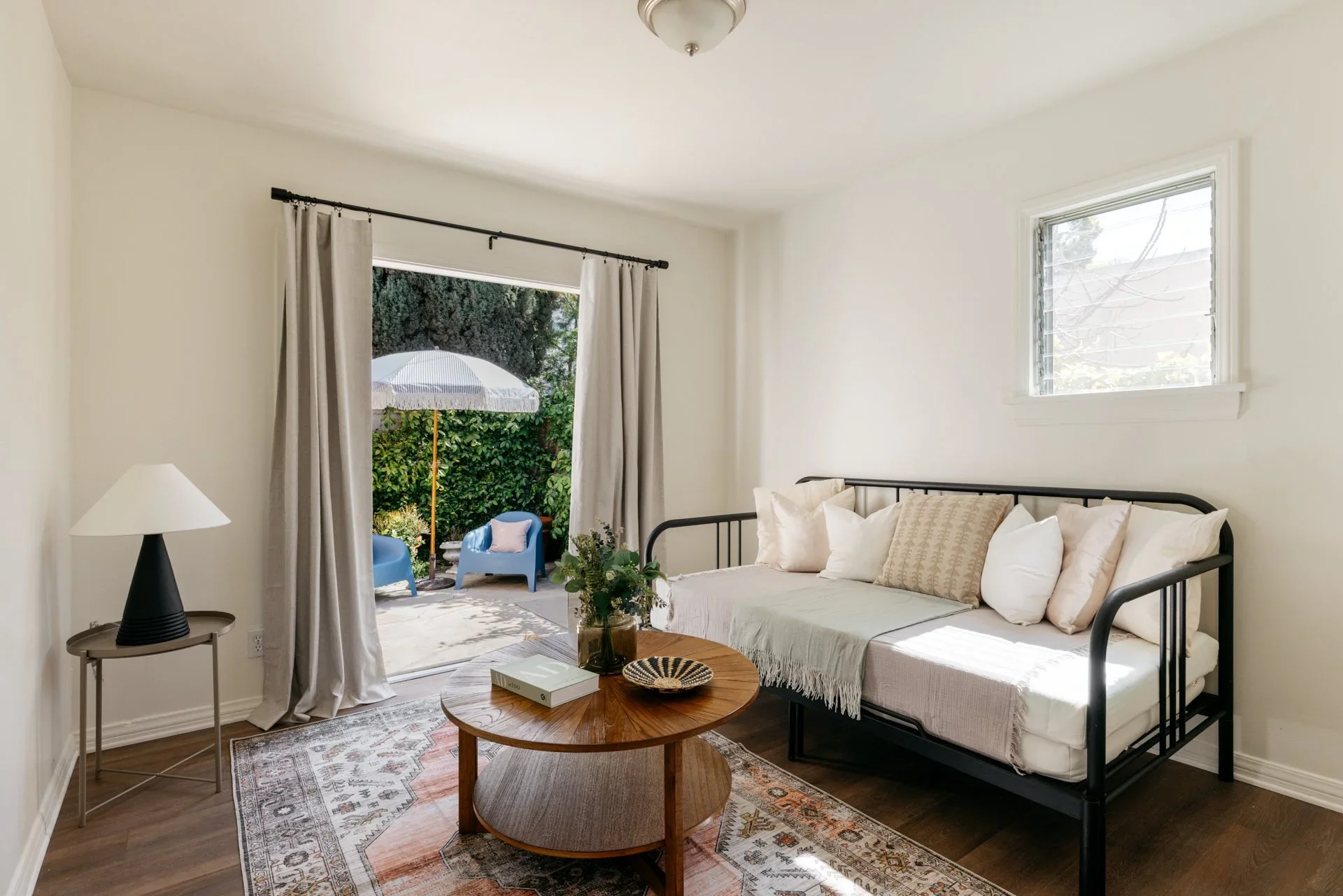 A cozy living room with a black frame daybed covered with white and beige pillows, a small wooden coffee table with books and a green plant, a patterned area rug, a black side table with a white lampshade, and a sliding glass door leading to a small outdoor patio with blue chairs and a white umbrella, letting in natural light.