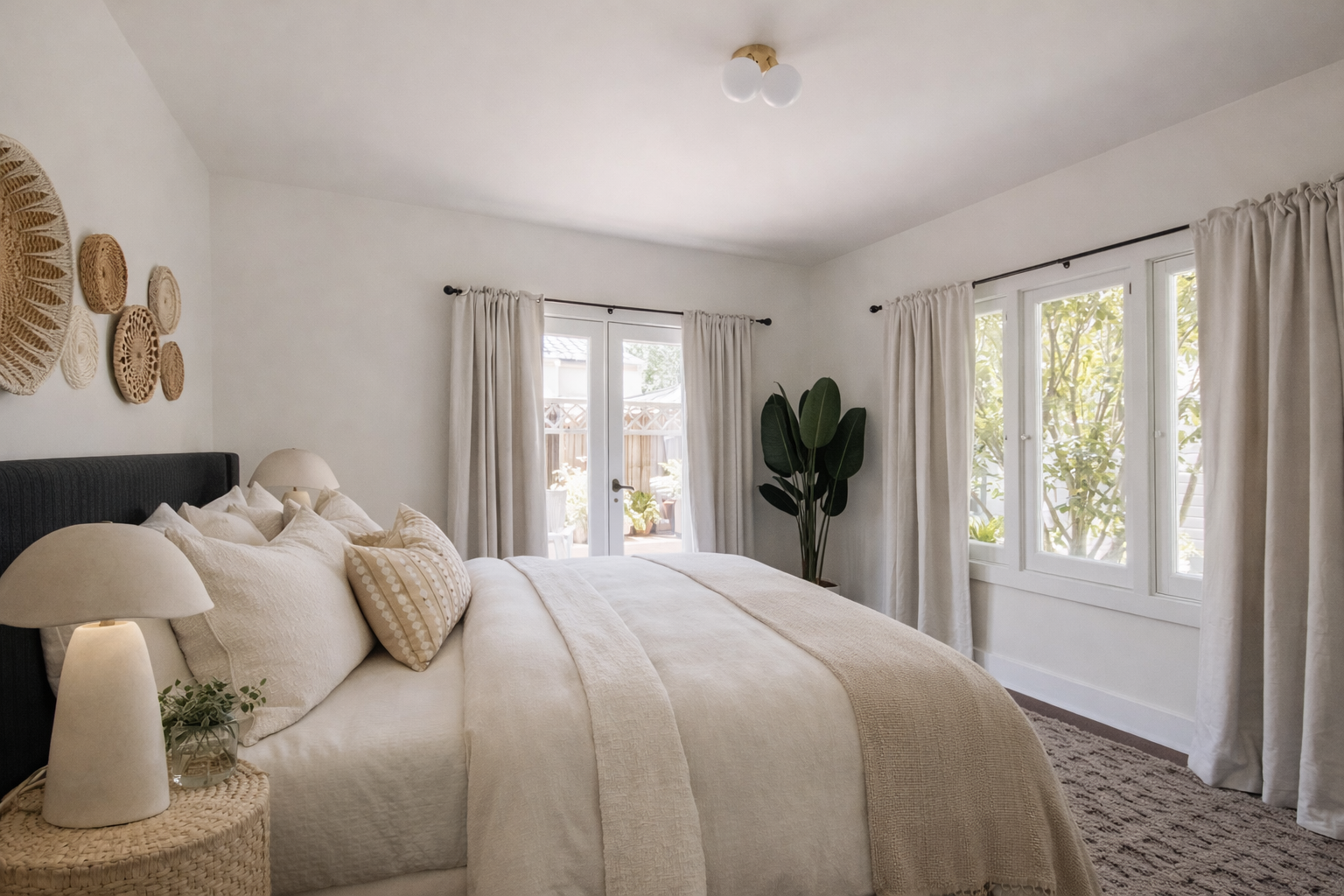 A cozy bedroom with a large bed dressed in white linens and beige pillows, two bedside lamps, a large potted plant, large windows with white curtains, and wall decor of woven baskets.
