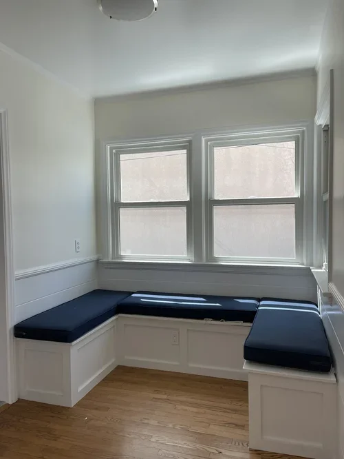 A built-in window seat with navy cushions in a small room with two large windows, white walls, and hardwood floors.