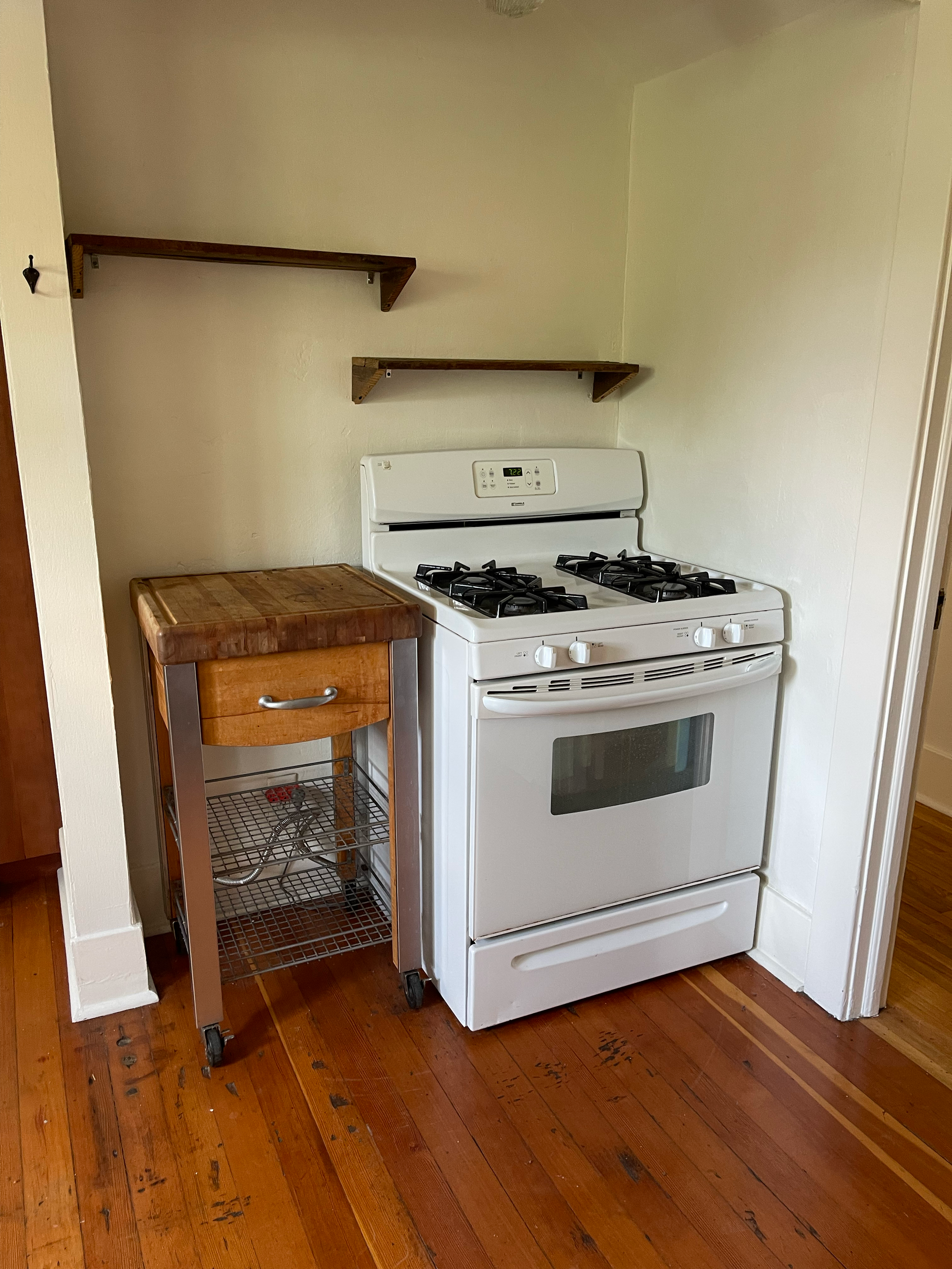 A white gas stove with four burners and an oven, next to a small wooden and metal cart with storage racks, in a corner of a kitchen with wooden flooring and two wooden shelves above.