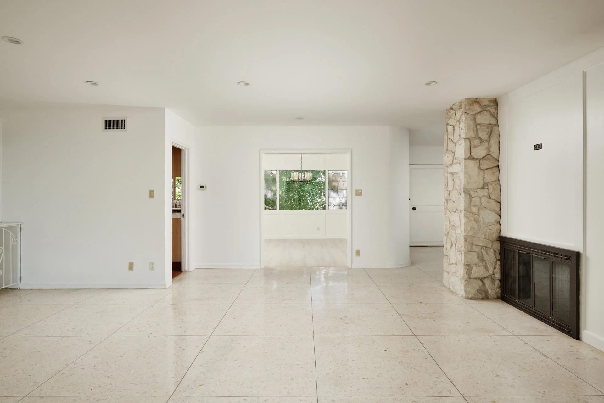 Empty living room with white walls, beige tile flooring, a stone fireplace, and a large window revealing greenery outside.