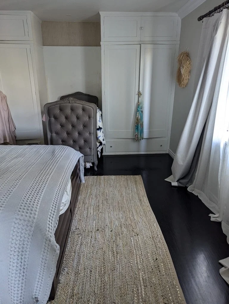 A bedroom with a white wardrobe, a beige tufted headboard on the bed, a cream-colored rug on dark hardwood floor, white curtains on a window, and a wall hanging on the wall.