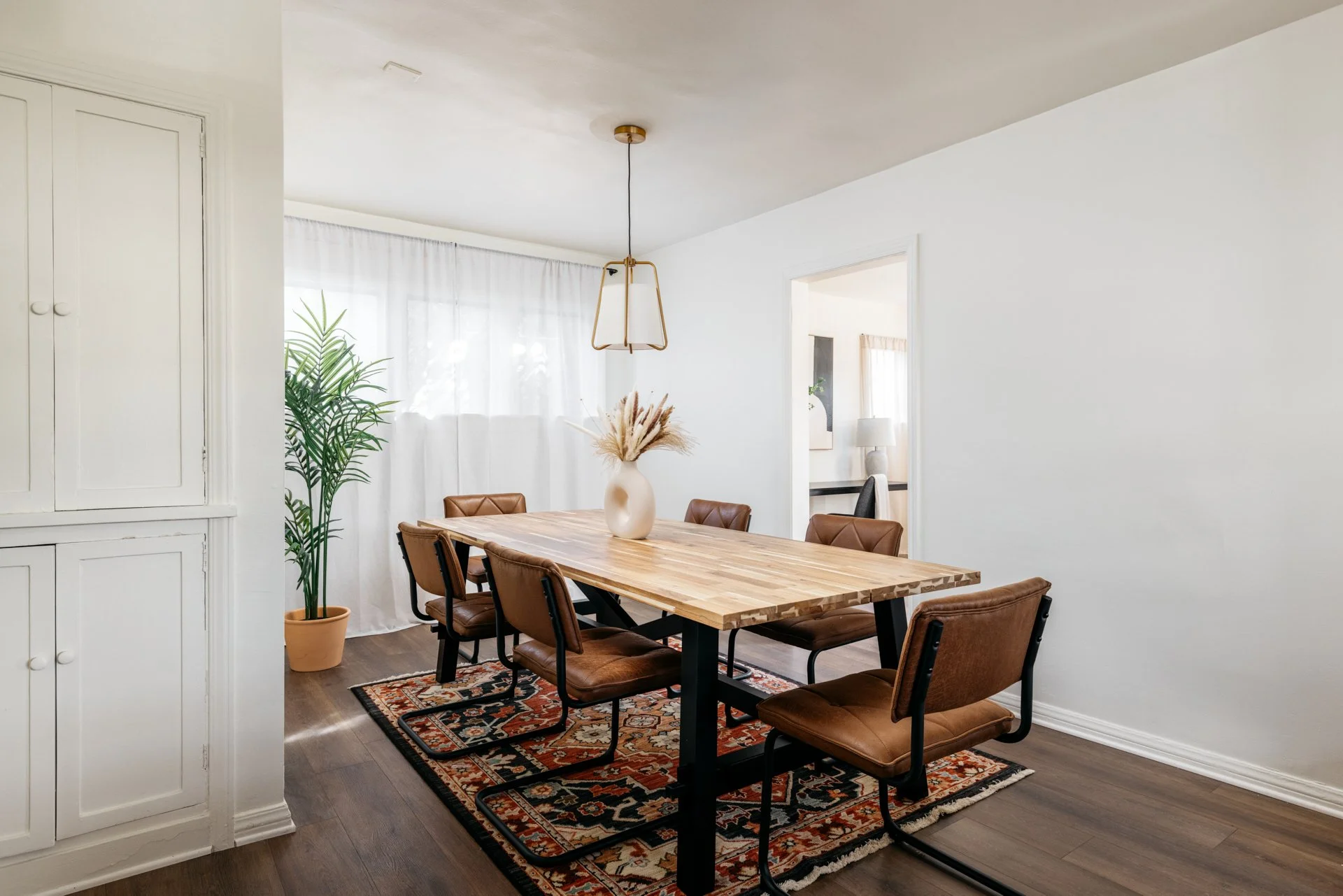 Dining room with a wooden table, six brown chairs, a vase with dried flowers, a potted plant, a patterned rug, and a window with white curtains.