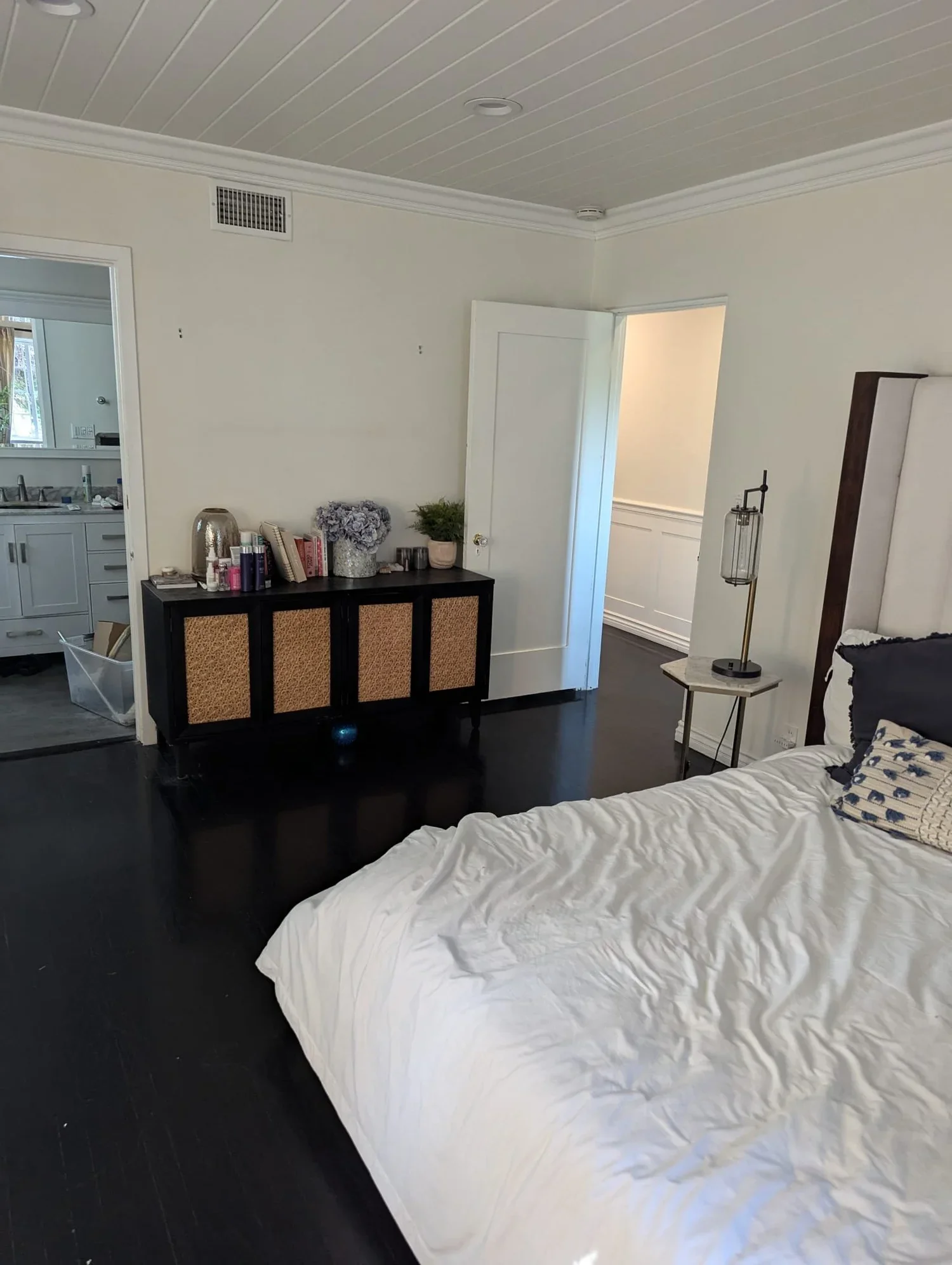 A bedroom with a bed, black hardwood floor, dresser with decor and books, nightstand with lamp, and open door leading to another room.