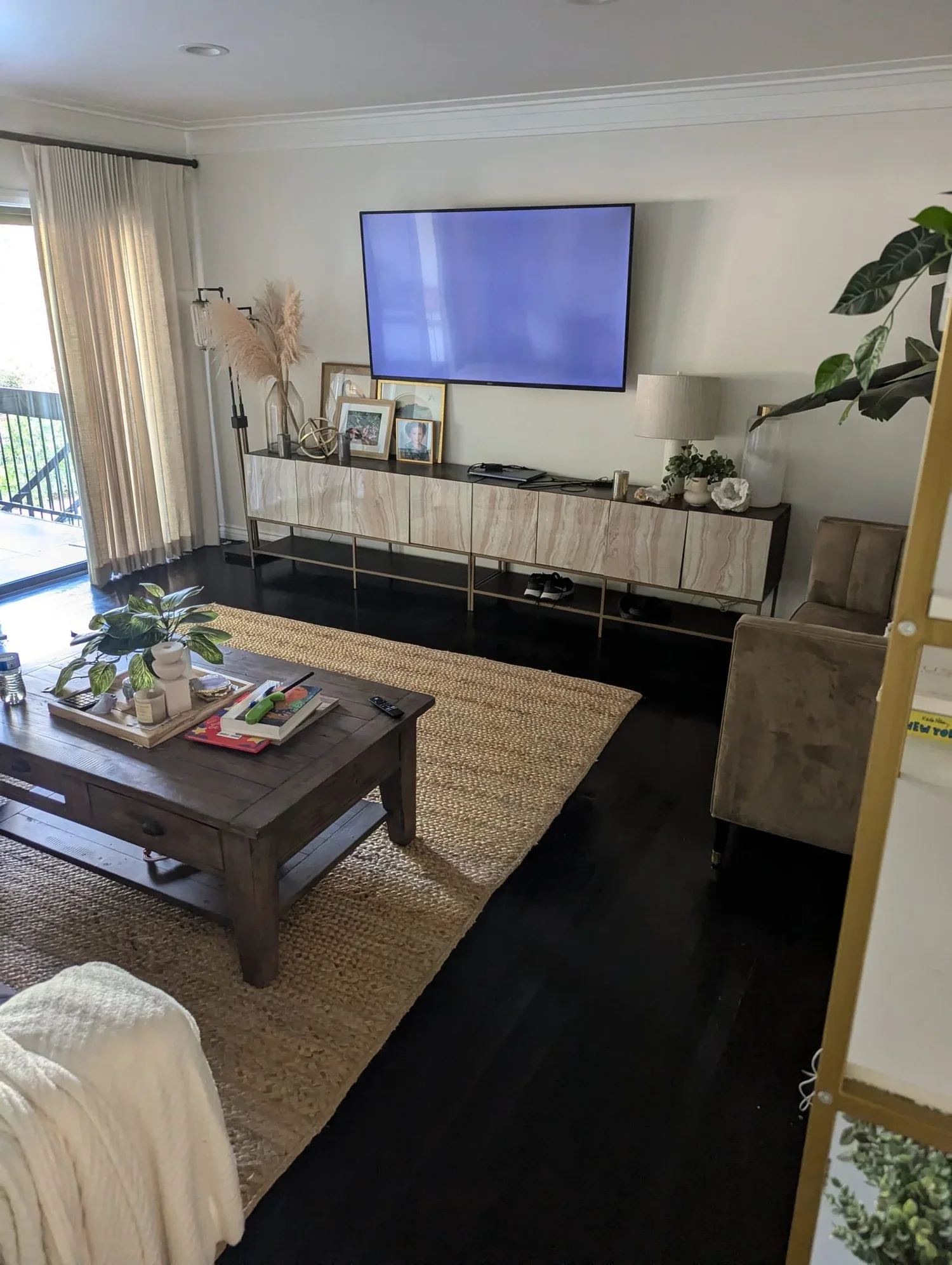 Living room with a mounted flat-screen TV, a decorative sideboard, a coffee table with plants and books, a sliding glass door with curtains leading outside, and a beige armchair.