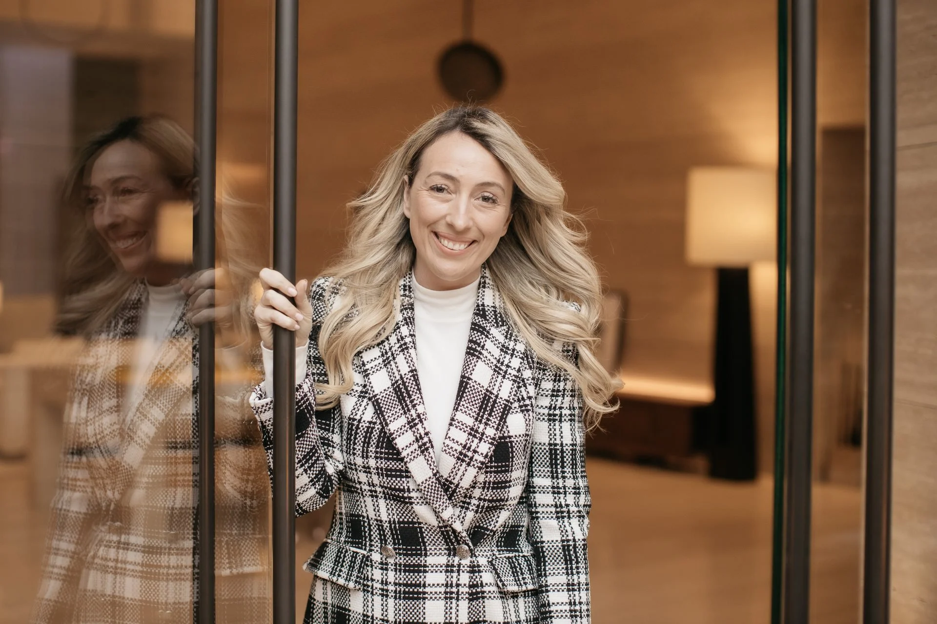 A woman with long blonde Hair smiling. She is holding onto elevator door frame. Inside elevator with wooden walls and a lamp visible in the background.