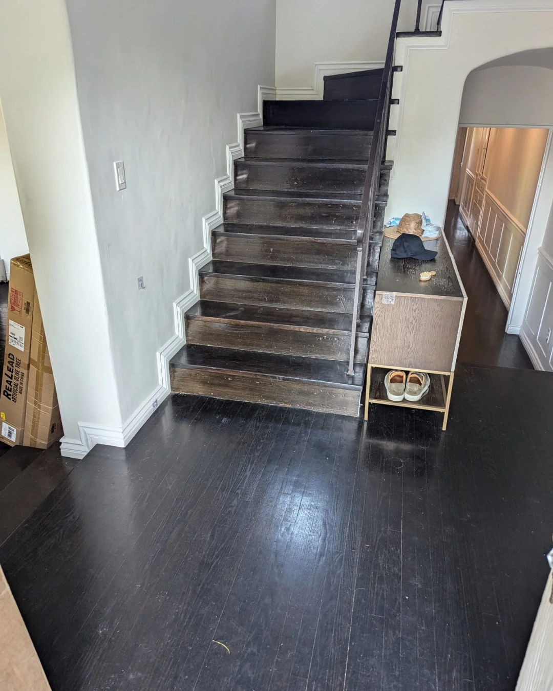 Interior image of a staircase with dark wooden steps and black handrail, adjacent to a small wooden storage unit with shoes underneath, located at the entrance of a home with white walls and dark hardwood floors.