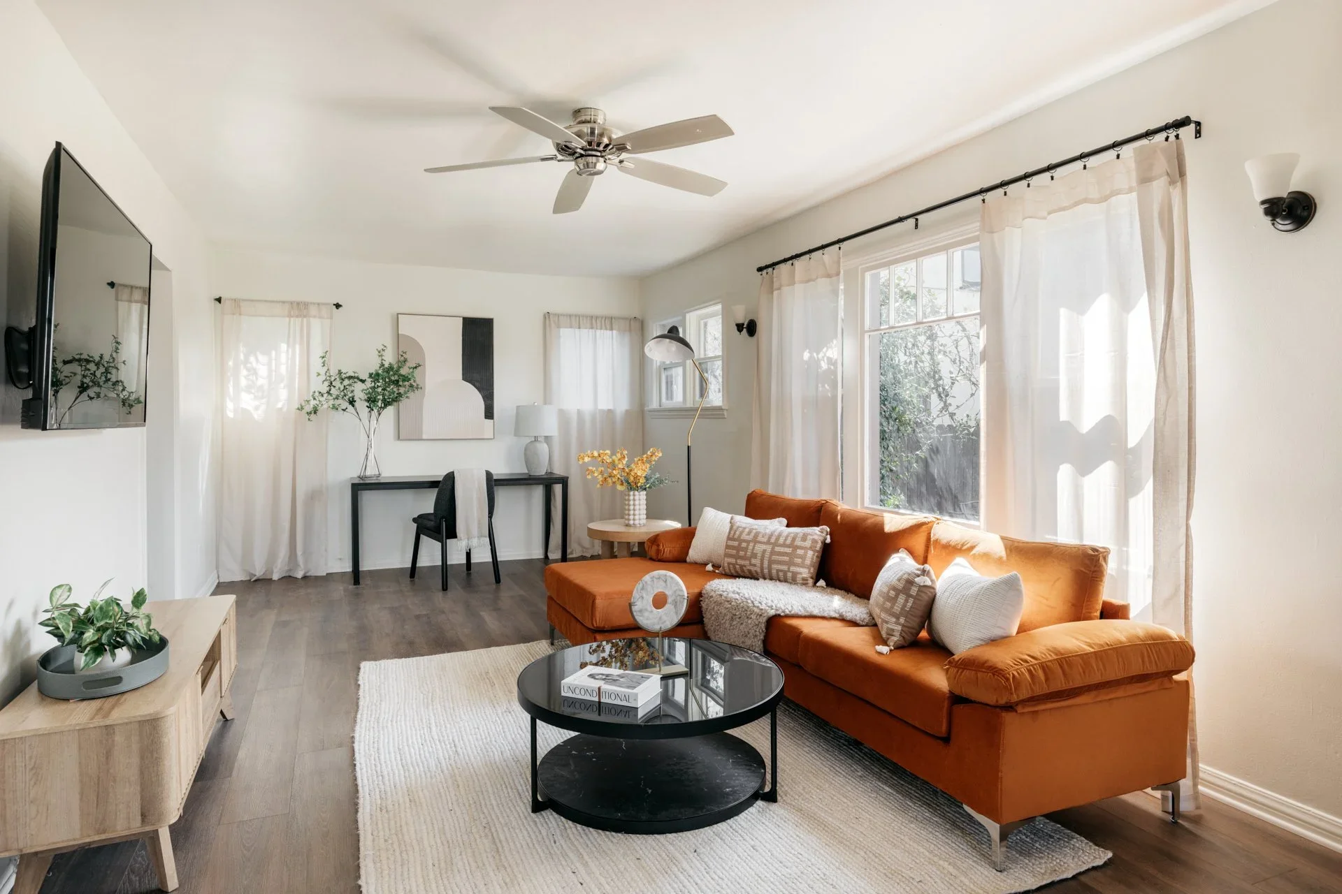 Bright living room with orange sofa, black round coffee table, white rug, windows with sheer curtains, artwork on white walls, and black decor accents.