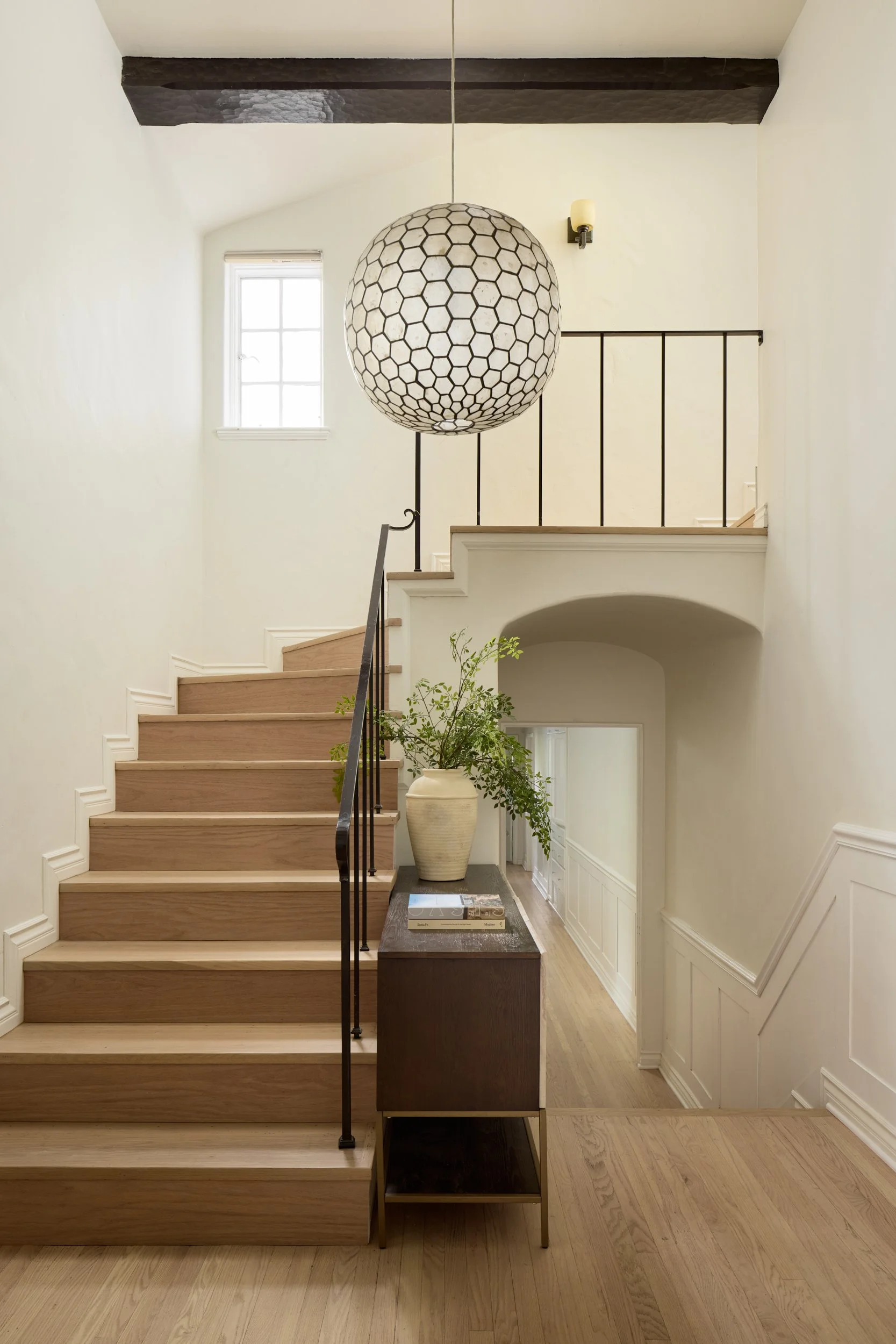 Interior of a bright, modern home entryway with wooden staircase, large hanging pendant light with honeycomb pattern, white walls, black metal railing, a small table with books and a large decorative vase with green foliage, and a small window near the ceiling.