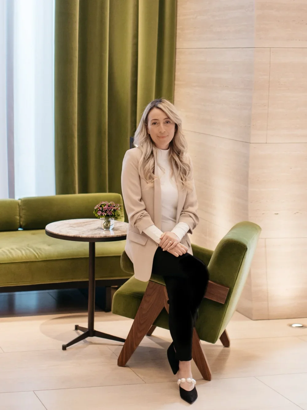 A woman with long blonde hair, wearing a beige blazer, white top, and black pants, sitting on a green chair with wooden legs in a modern lounge. Behind her is a green sofa, a small round marble-top table with a flower arrangement, green curtains, and a beige wall.