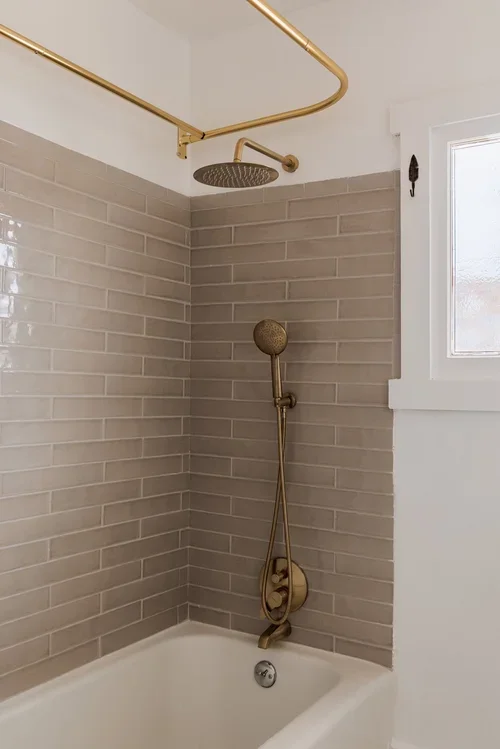Bathroom shower area with grey subway tile walls, a golden showerhead, handheld shower, and a window.