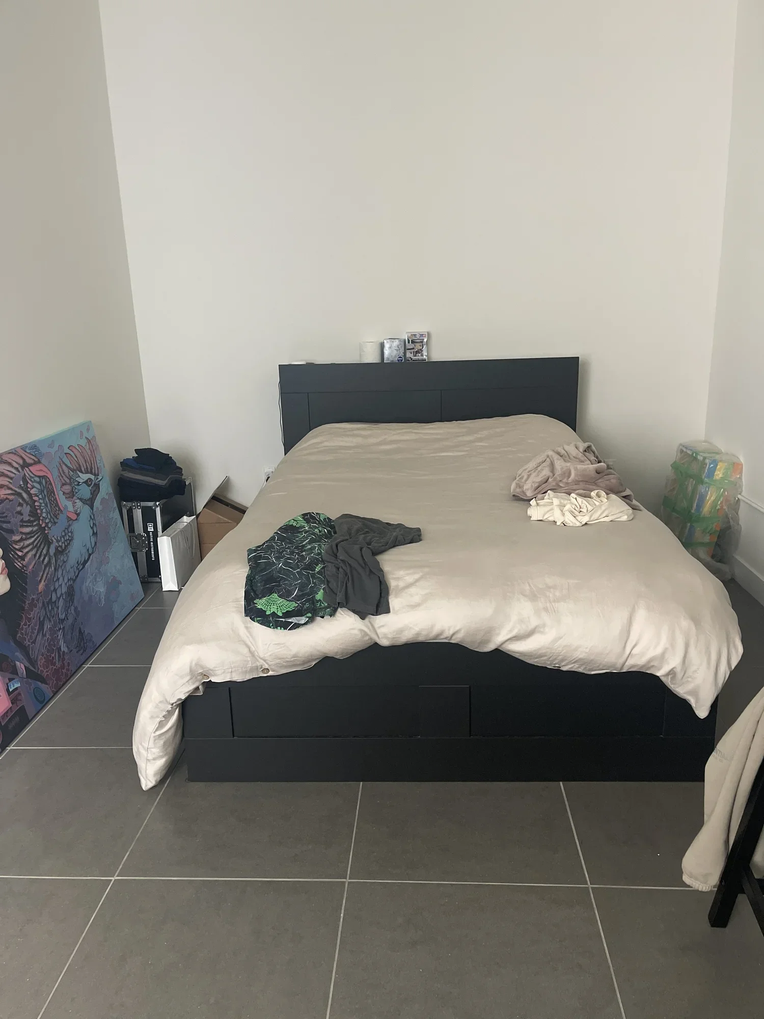 Unmade bed with beige sheets and pillows, various clothes on top, artwork leaning against the wall, and some boxes on the floor in a bedroom.