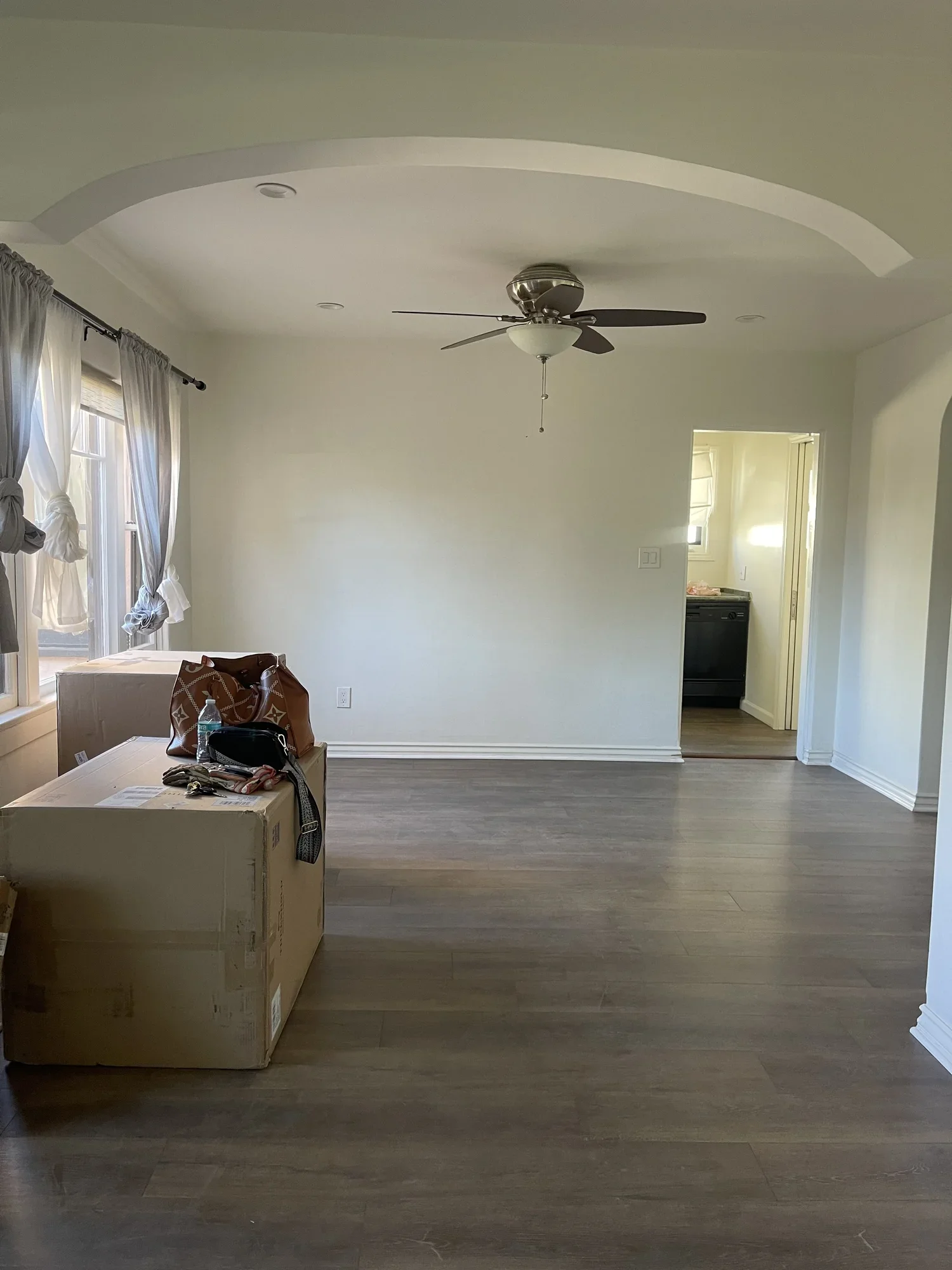 Empty living room with light-colored walls, a ceiling fan, and large windows with curtains. There are some cardboard boxes and personal items on a box near the window.