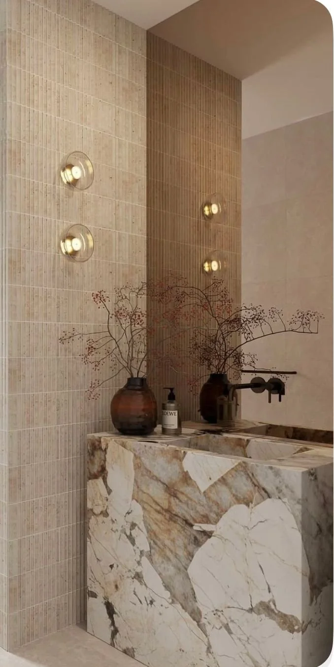 A modern bathroom with a marble vanity, two vases with dried branches, a soap dispenser, and a wall mirror with four round wall lights.
