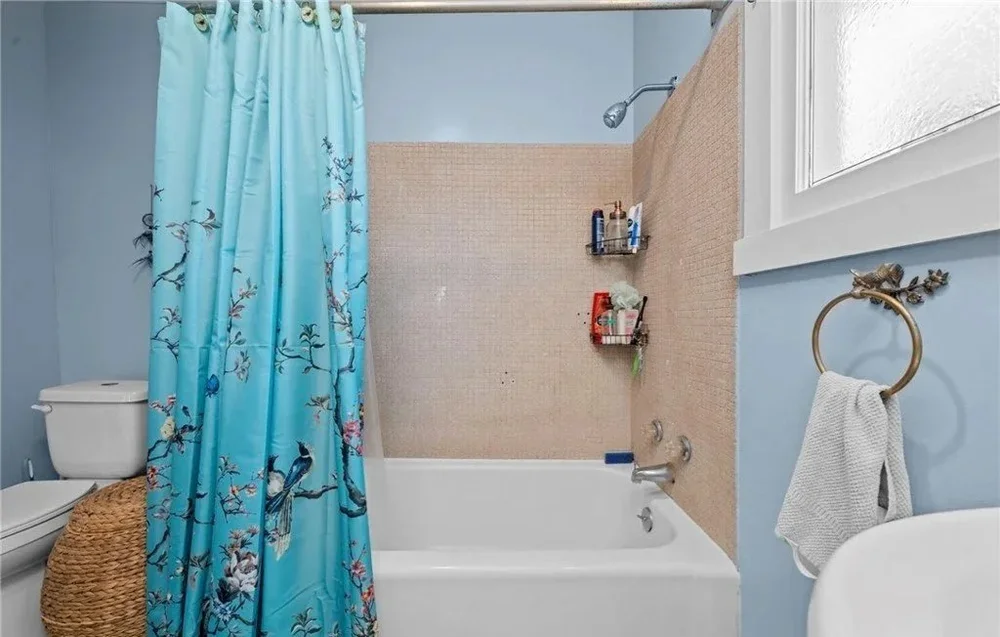 Bathroom with light blue walls, a bathtub with a beige tile surround, a blue shower curtain with birds and flowers, a white toilet, a wicker basket next to the toilet, a metal towel ring with a white towel, and windows with white framing.