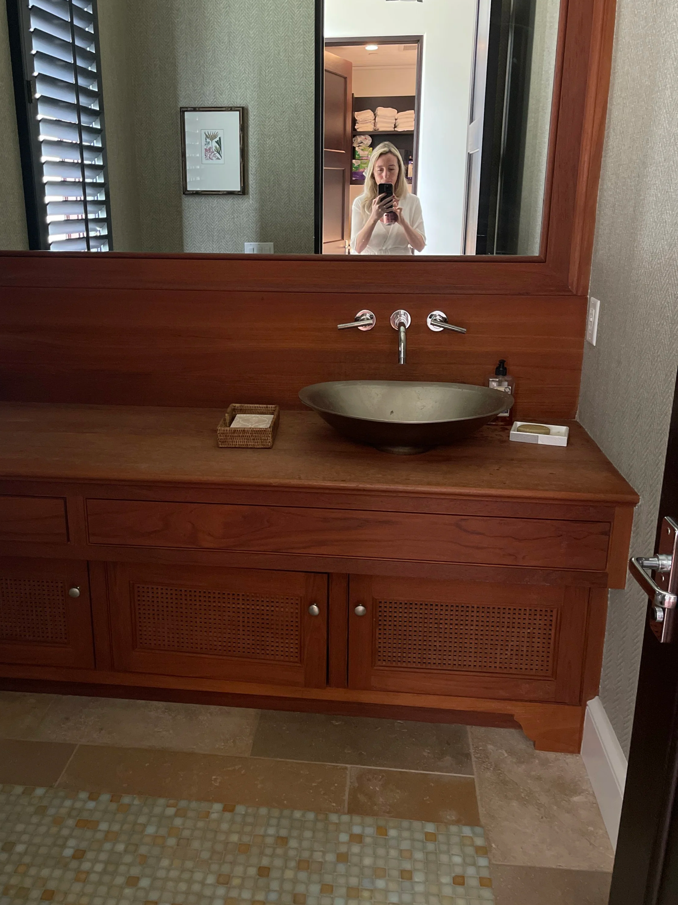 A bathroom vanity with a wooden cabinet, a ceramic vessel sink, and a large mirror reflecting a woman taking a photo with her phone, standing in what appears to be a bedroom or closet area with shelves of towels.