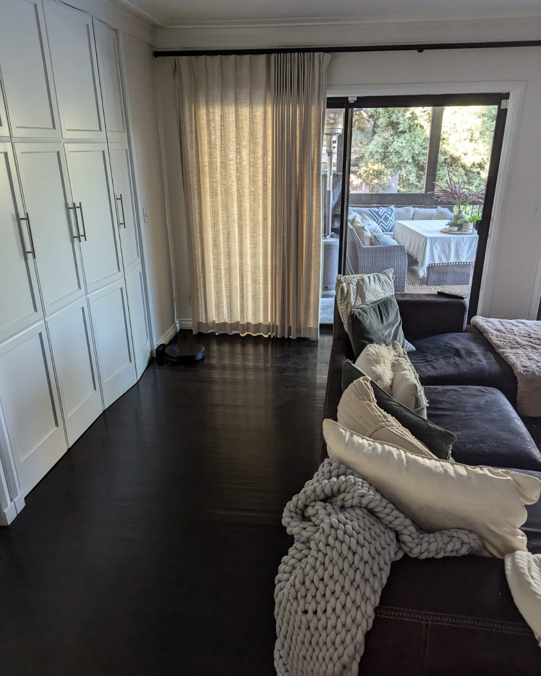 Living room with dark wood flooring, white built-in cabinets, a gray sectional sofa with pillows, and sliding glass doors leading to a screened-in porch with outdoor seating and trees outside.