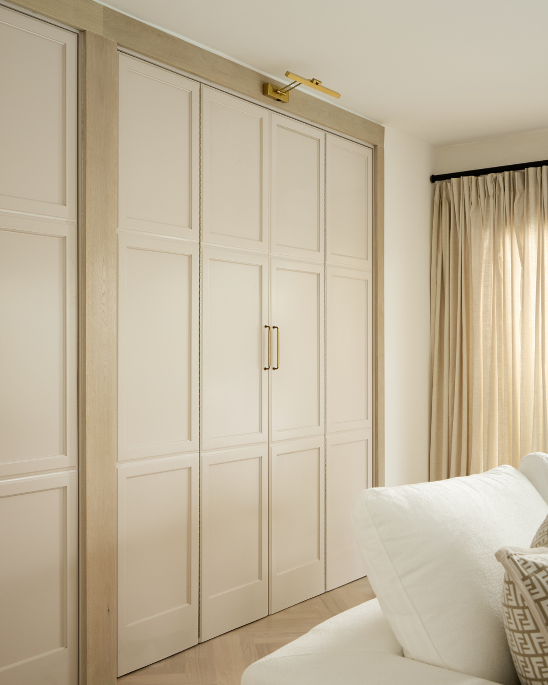 A built-in closet with paneled white doors, gold handles, and a wood trim, in a well-lit room with beige curtains and cream-colored furniture.