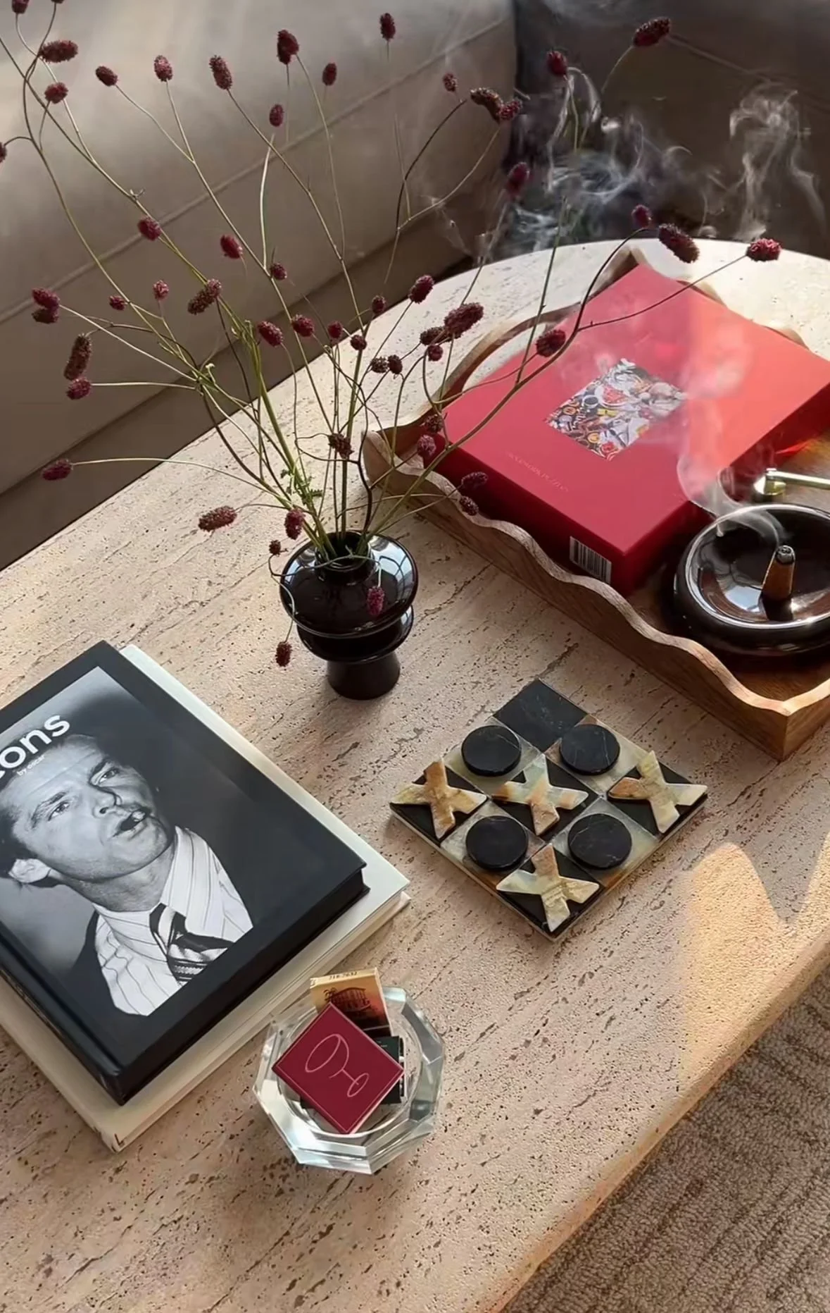 A table with a black vase holding dried wildflowers, a book with a black-and-white photo of a man, a glass dish with small matches and red matchboxes, a tray with a red item and a lit cigarette, and a chessboard with black and beige pieces.