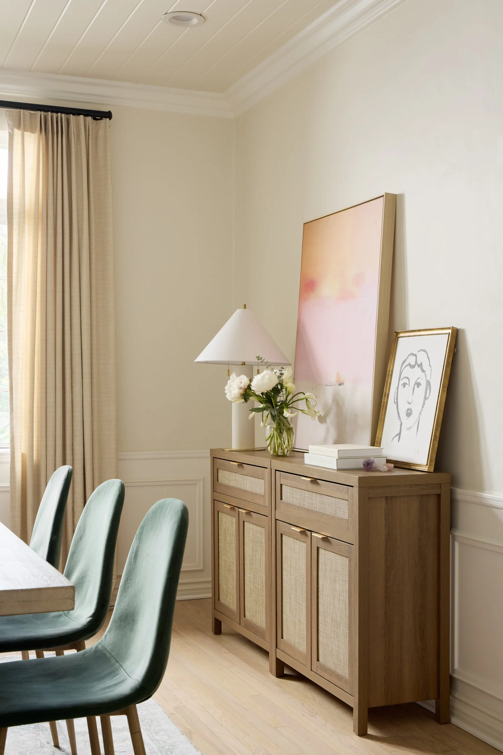 A wooden sideboard with a white lamp, a vase with white flowers, a framed line art portrait, and a large pink and orange abstract painting on a white wall in a dining room with cream curtains and light wood flooring.