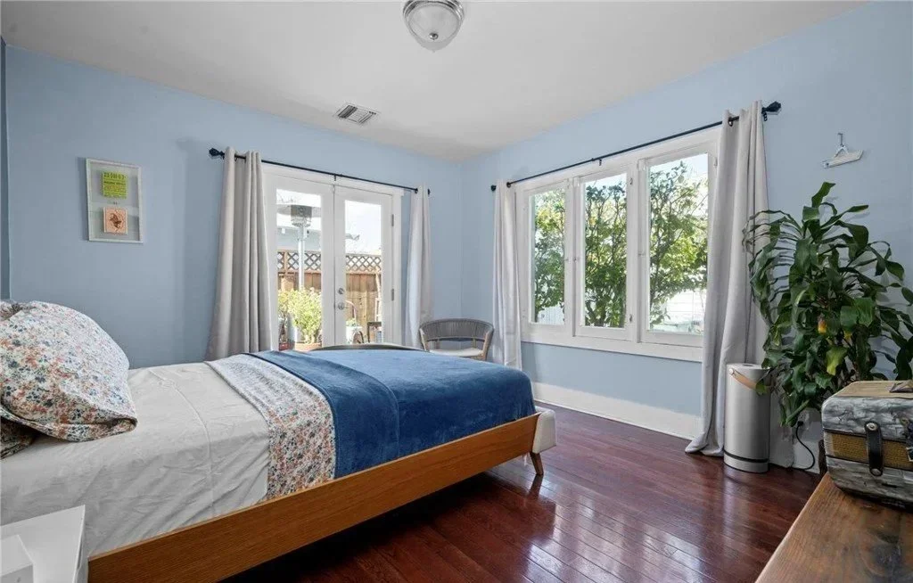 A bedroom with light blue walls, a bed with floral and white bedding, a dark blue blanket, and wooden flooring. There are large windows with white curtains, a chair, a tall plant, and a suitcase in the room.