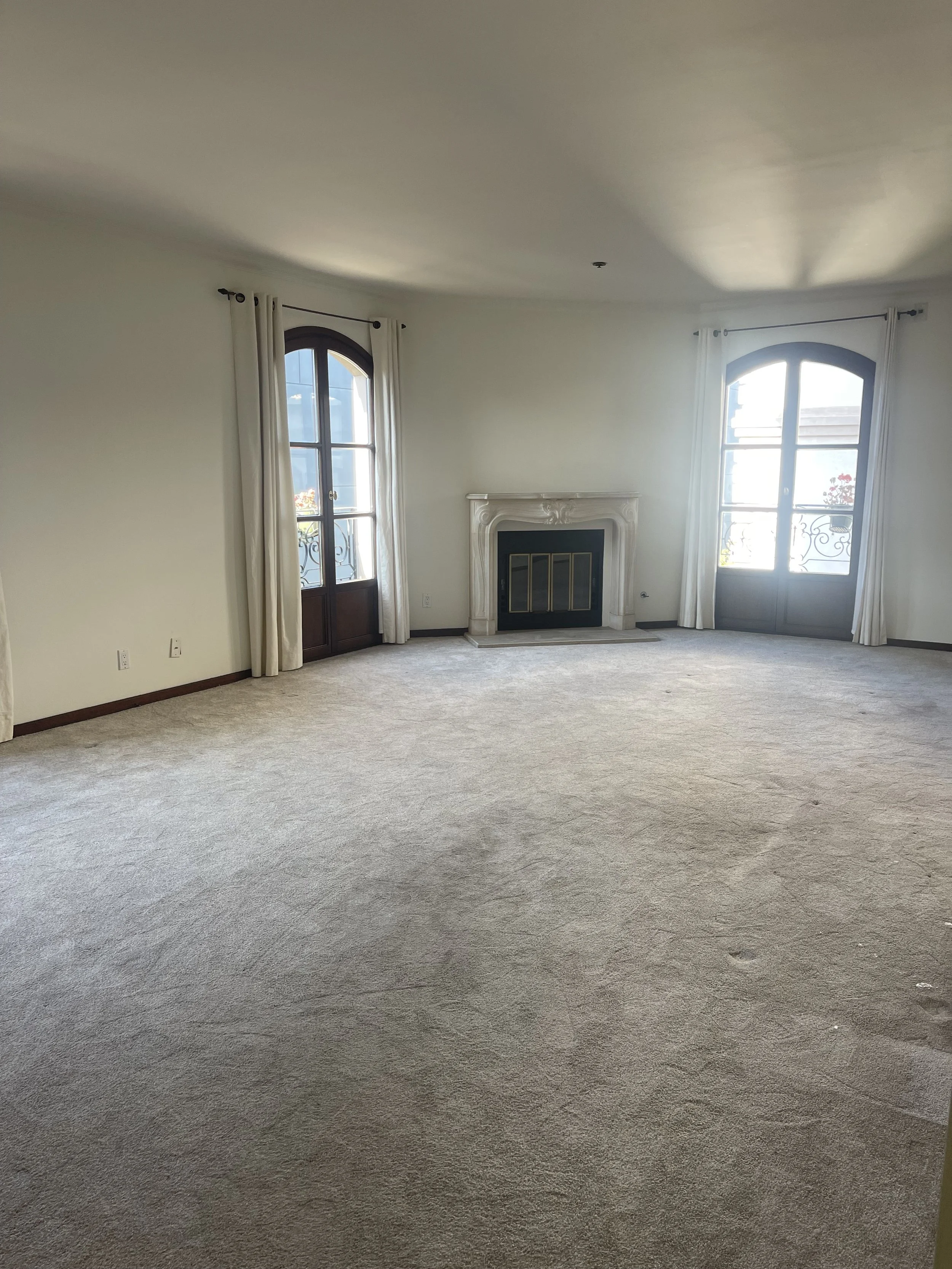 Empty living room with beige carpet, a fireplace, and two large windows with white curtains.