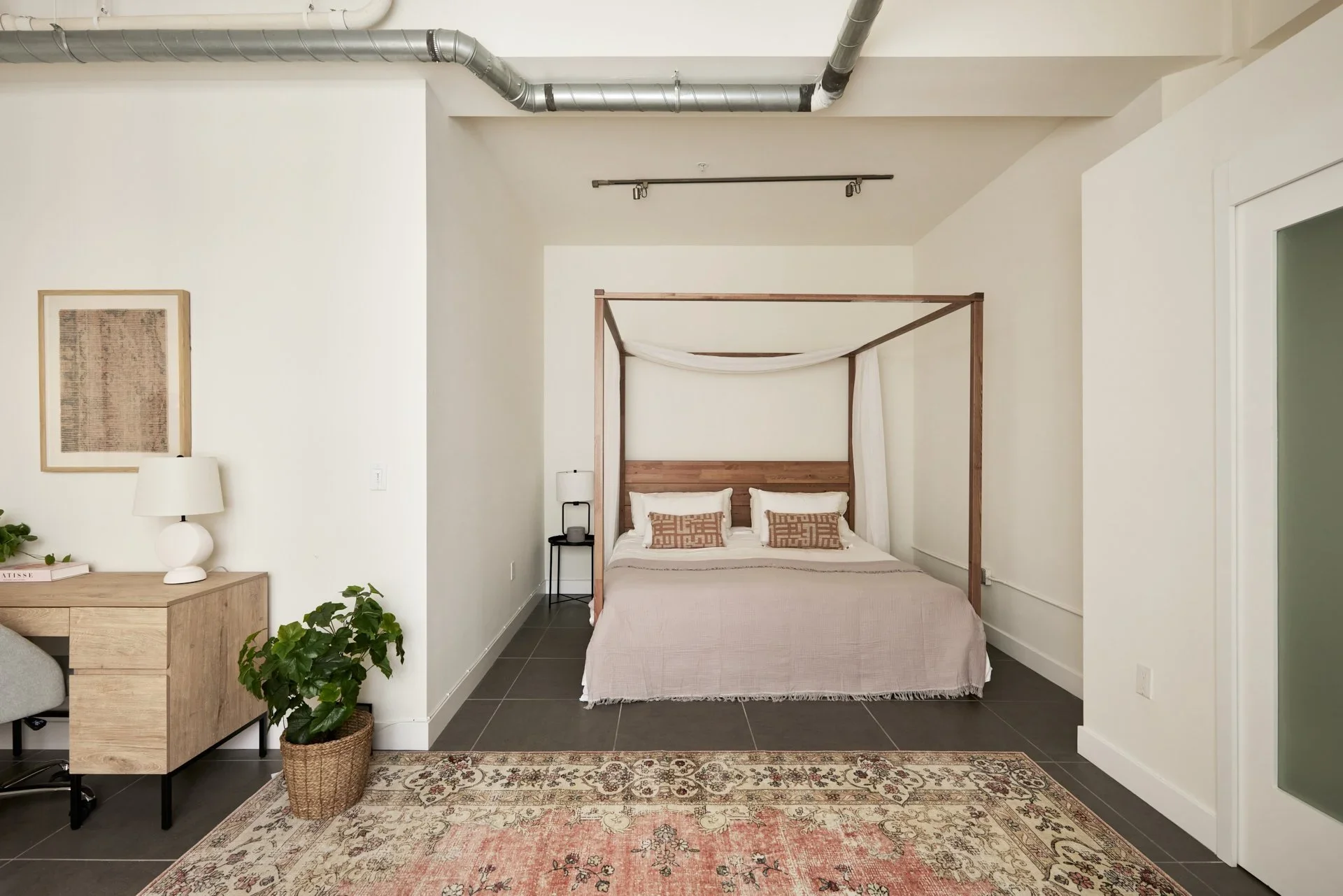 A cozy bedroom featuring a four-poster bed with white curtains and throw pillows. The room contains a nightstand with a lamp, a framed artwork on the wall, and a colorful area rug on gray tiled floor. There's a desk with a lamp and a potted plant nearby.