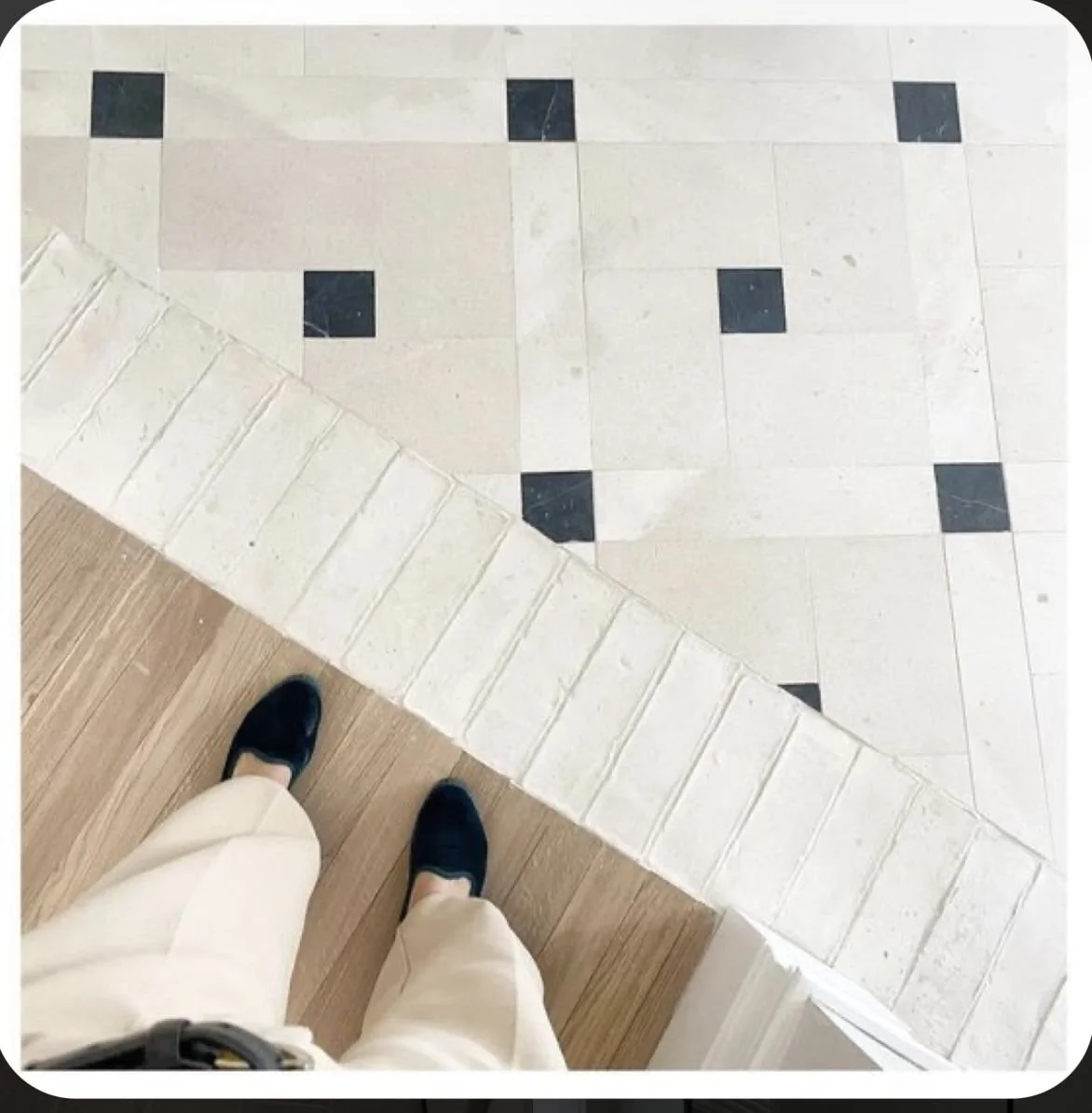 View of a person standing on a wooden floor, looking down at tile flooring with black and white tiles, with a low wall or step to the left.
