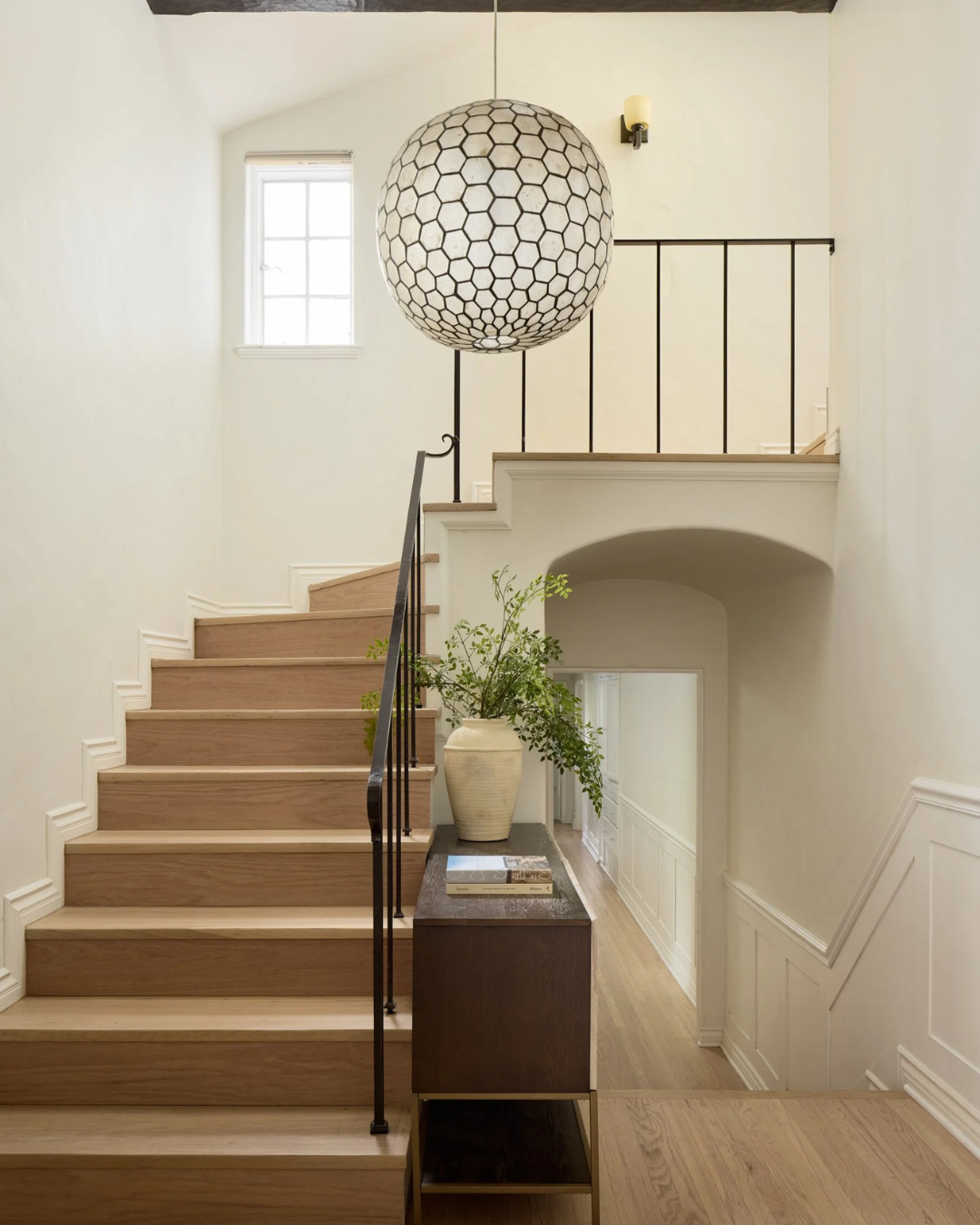 Interior view of a staircase with wooden steps, black railings, a large hanging geometric pendant light, a window, a side table with a potted plant and magazines, and cream-colored walls with white molding.