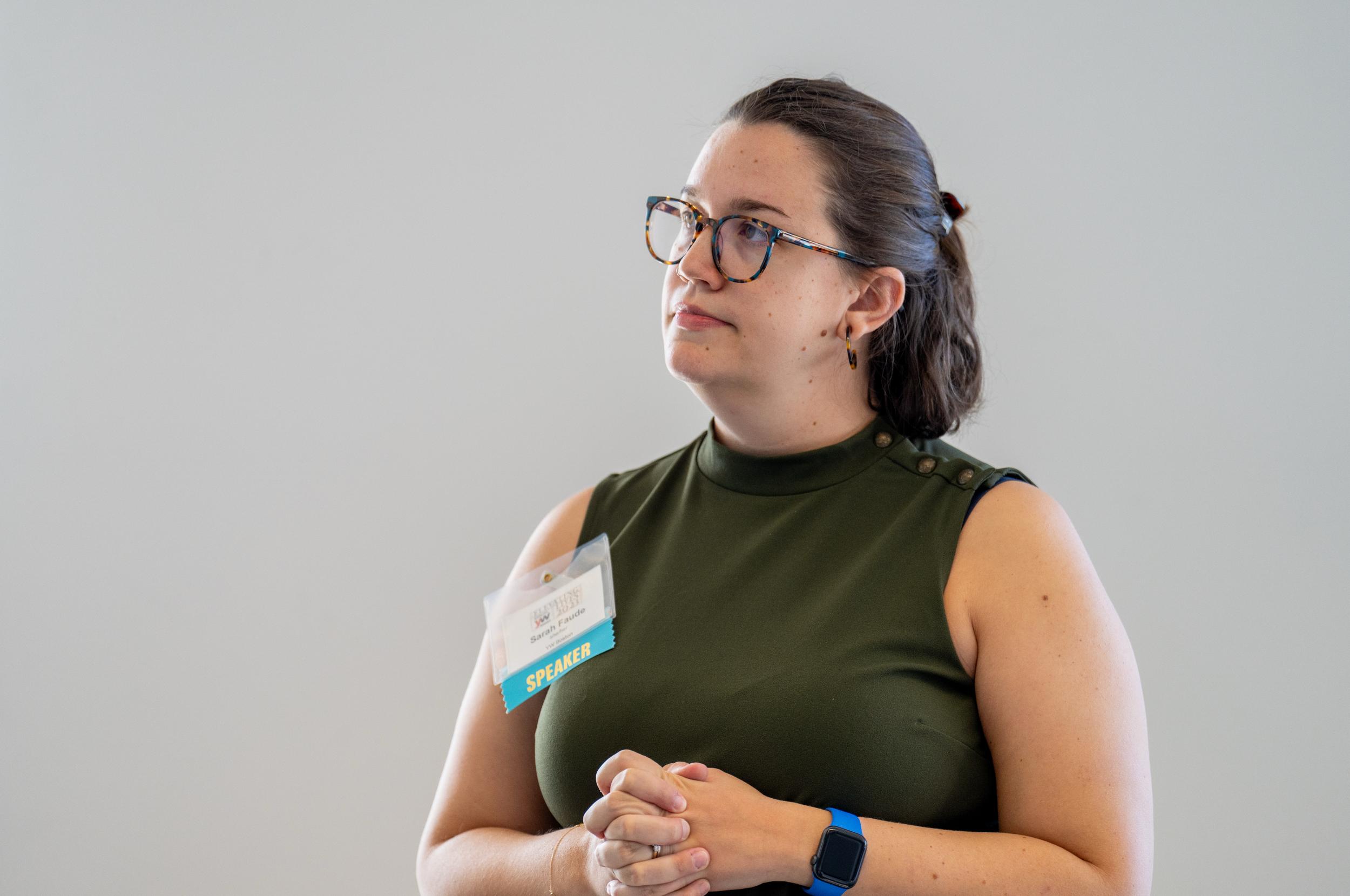 A woman with glasses standing with her hands clasped, wearing a sleeveless green top and a speaker badge, in front of a plain background.